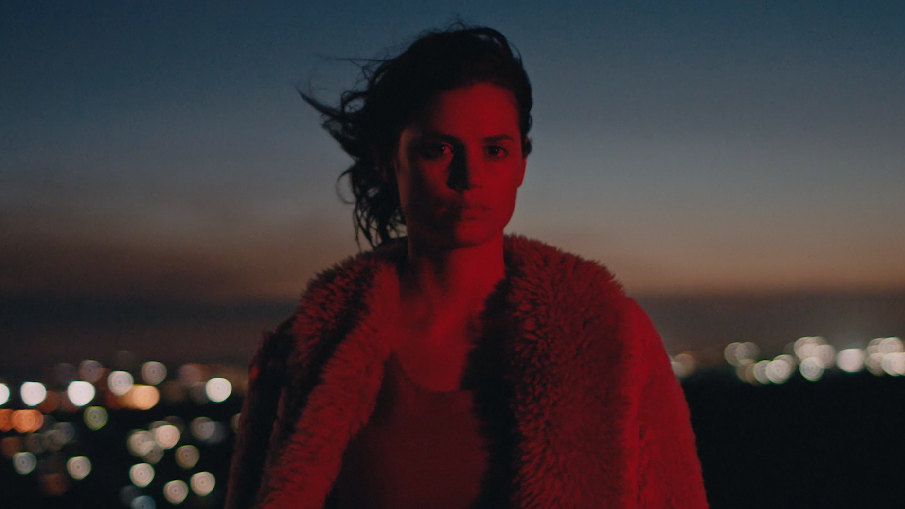 A woman with short hair stands outdoors at dusk, illuminated by red light, with city lights blurred in the background.