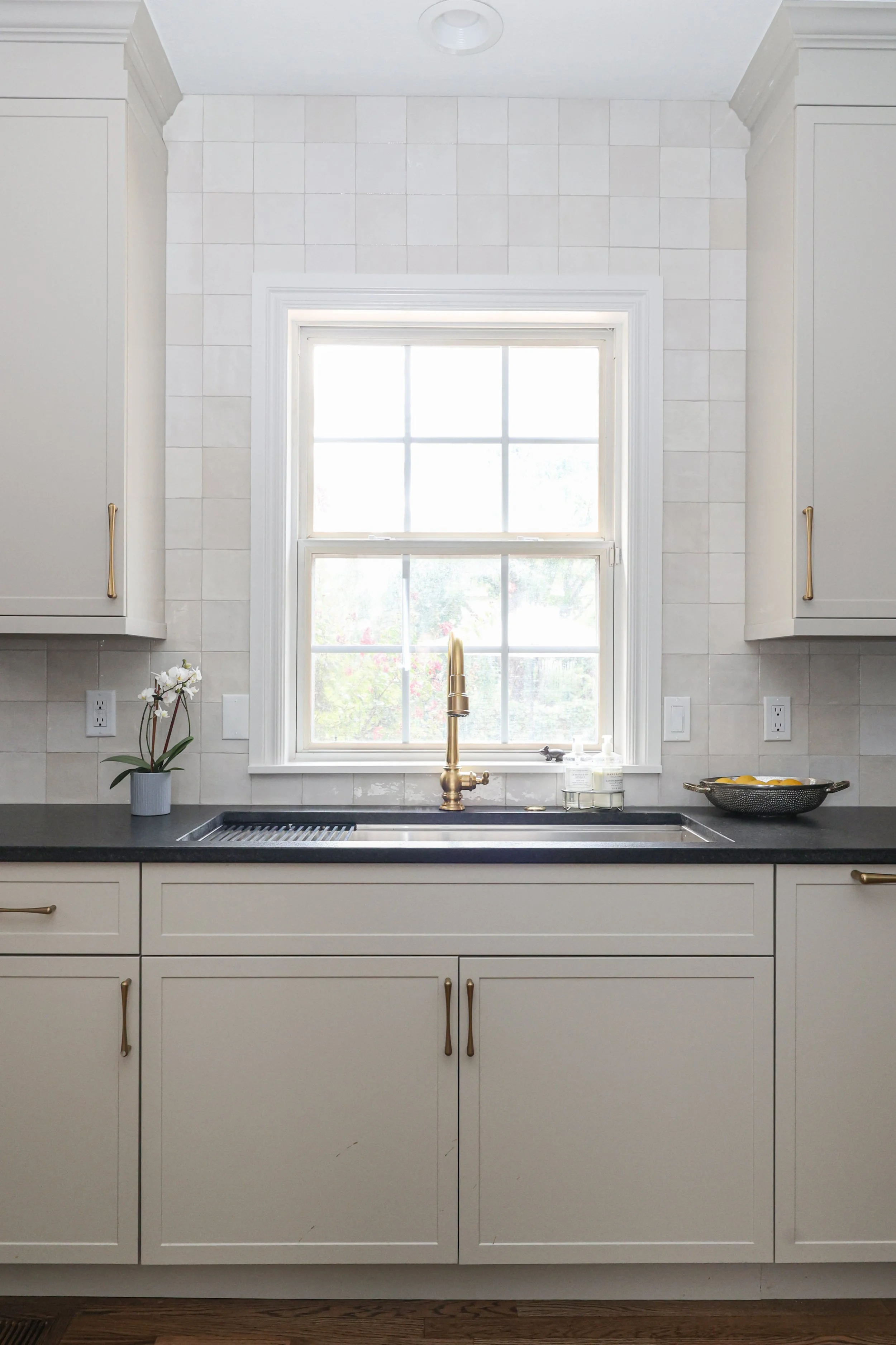 A kitchen sink area with white cabinetry, a black countertop, and a large window above the sink. There is a gold faucet, a potted orchid on the left, and a bowl of lemons on the right.