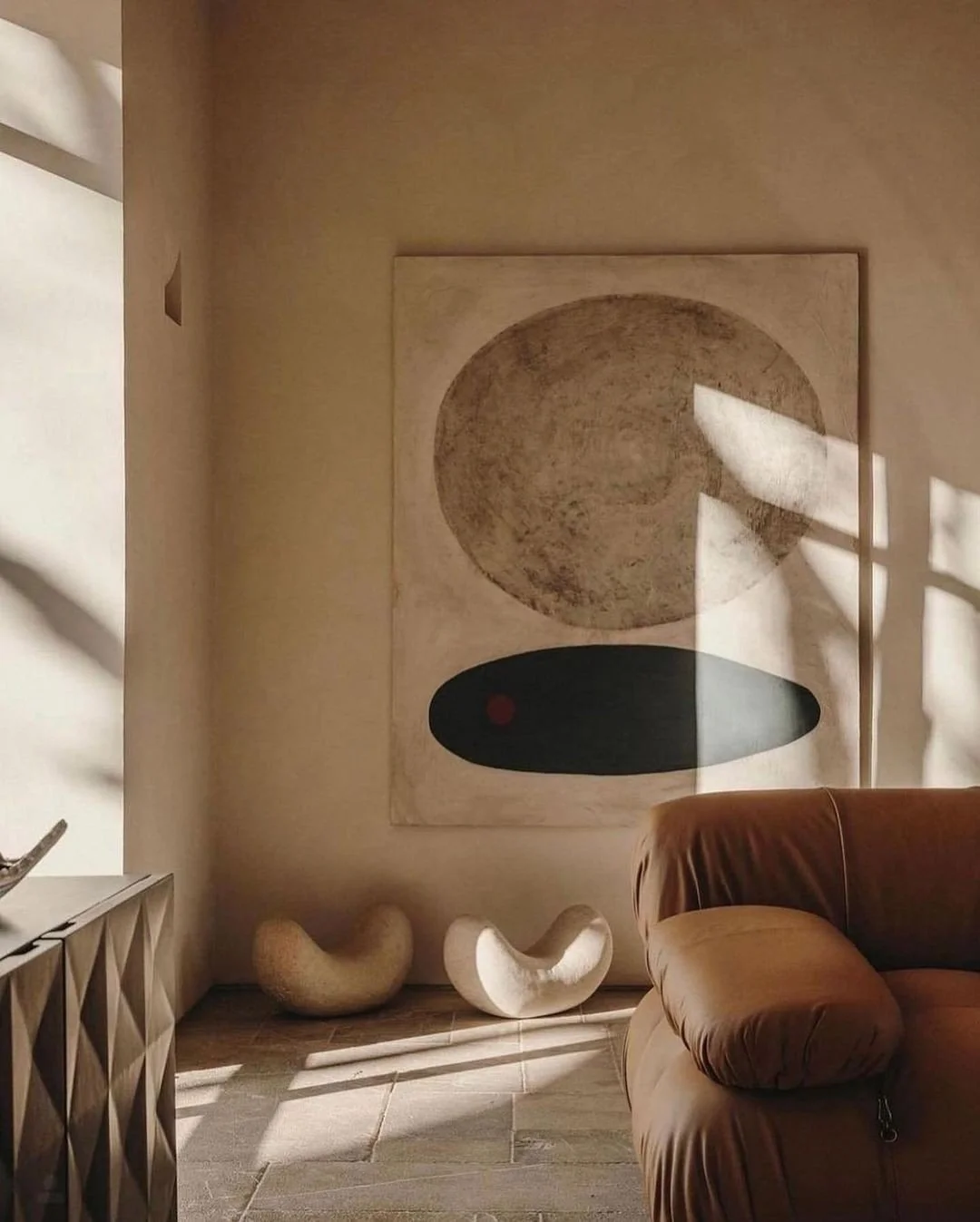 Sunlight creates shadow patterns on a beige wall with modern abstract artwork featuring circles and ovals. Two sculptural benches sit on the floor below the artwork. A brown sofa is partially visible on the right side.