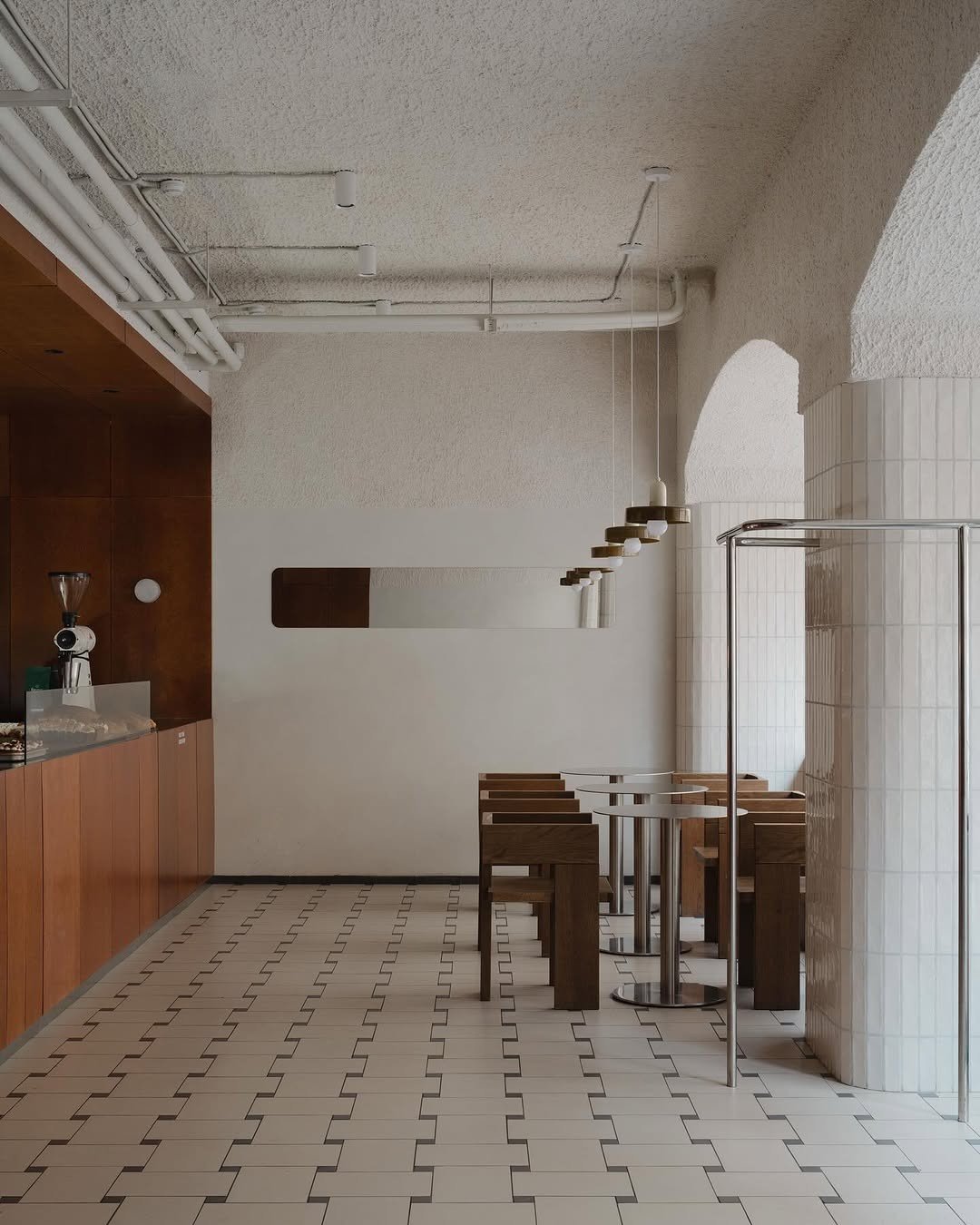 Empty café with wooden tables and benches, white tiled floor, and minimalist pendant lighting.