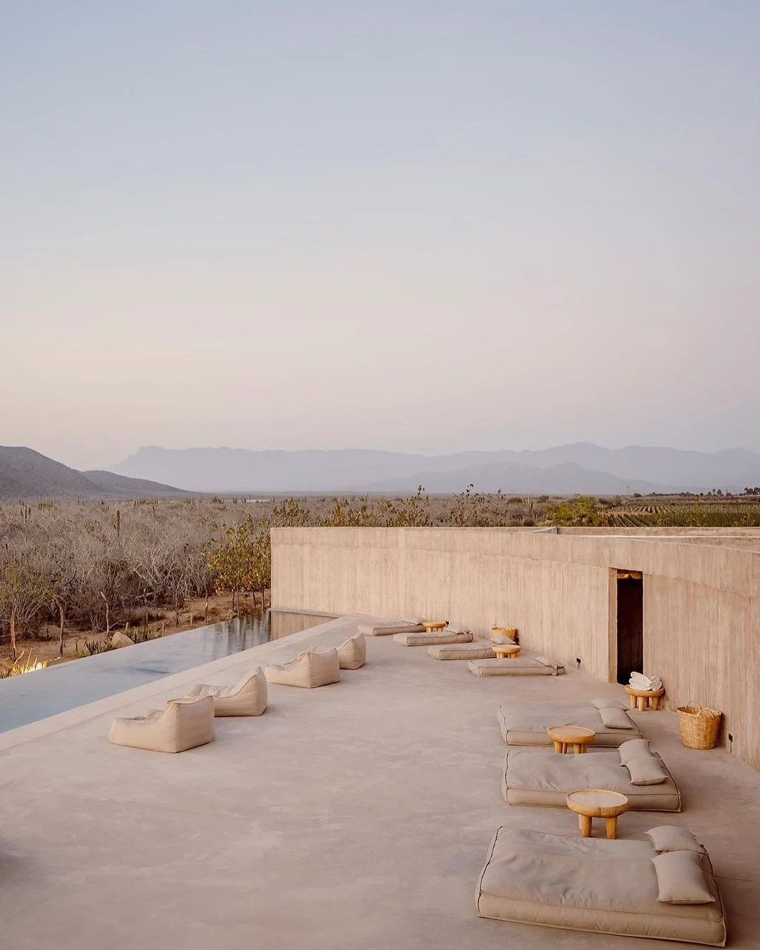 Outdoor terrace with beige cushions and small round tables, overlooking a desert landscape with mountains in the distance, under a clear sky.