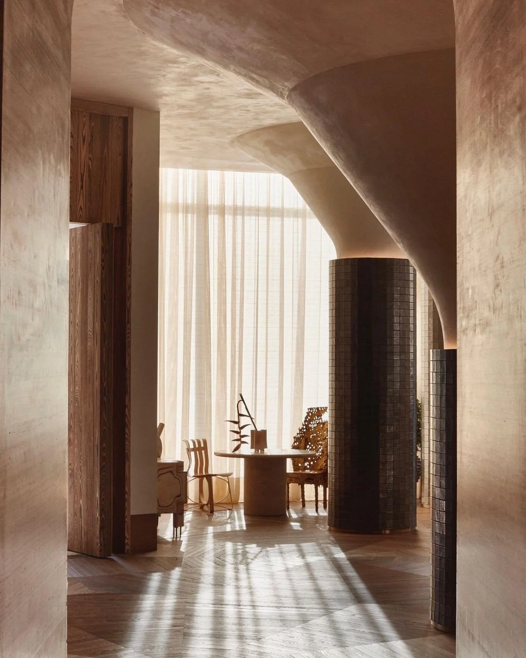 Elegant interior space with natural light, featuring a round table, chairs, decorative sculptures, and columns with textured patterns.