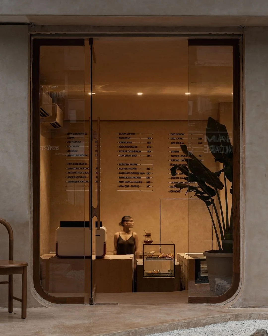 View through a glass window of a coffee shop interior with a woman behind the counter, a menu on the wall, a large potted plant to the right, and some baked goods displayed on a glass case.