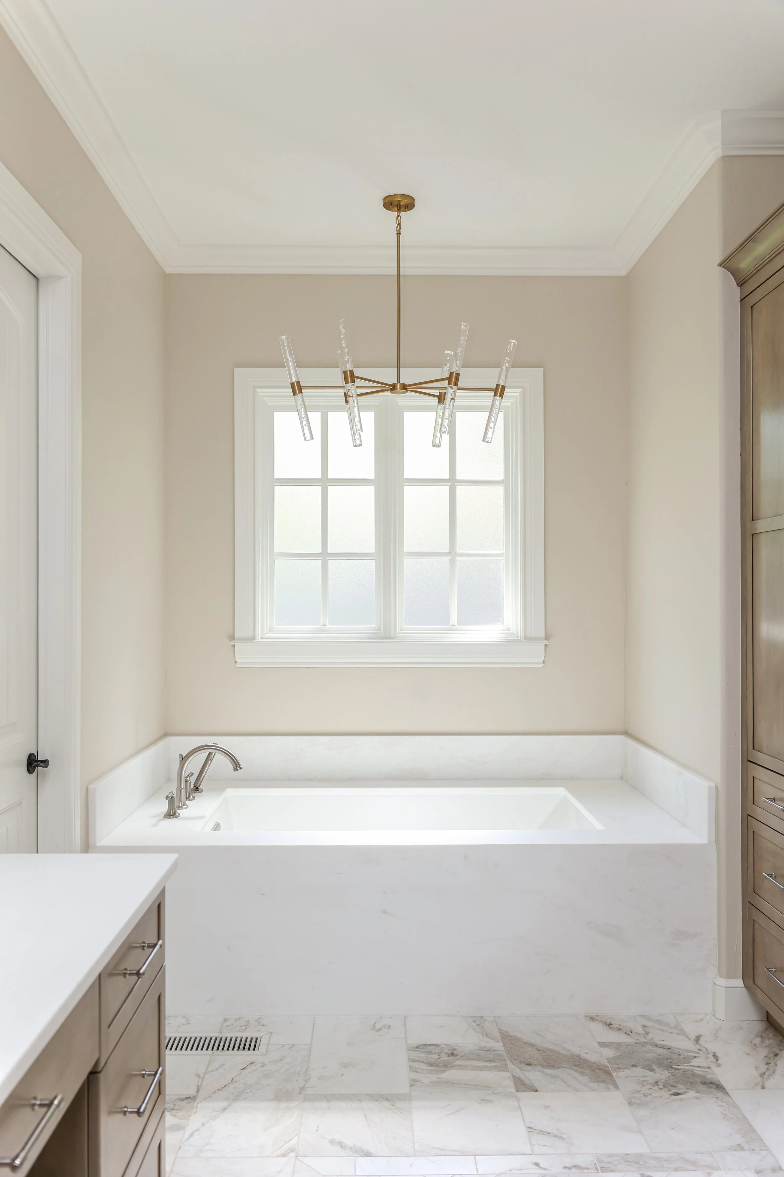 Modern bathroom featuring a built-in bathtub in white marble, a window behind the tub, beige walls, a brass chandelier with glass rods, and wooden cabinets.