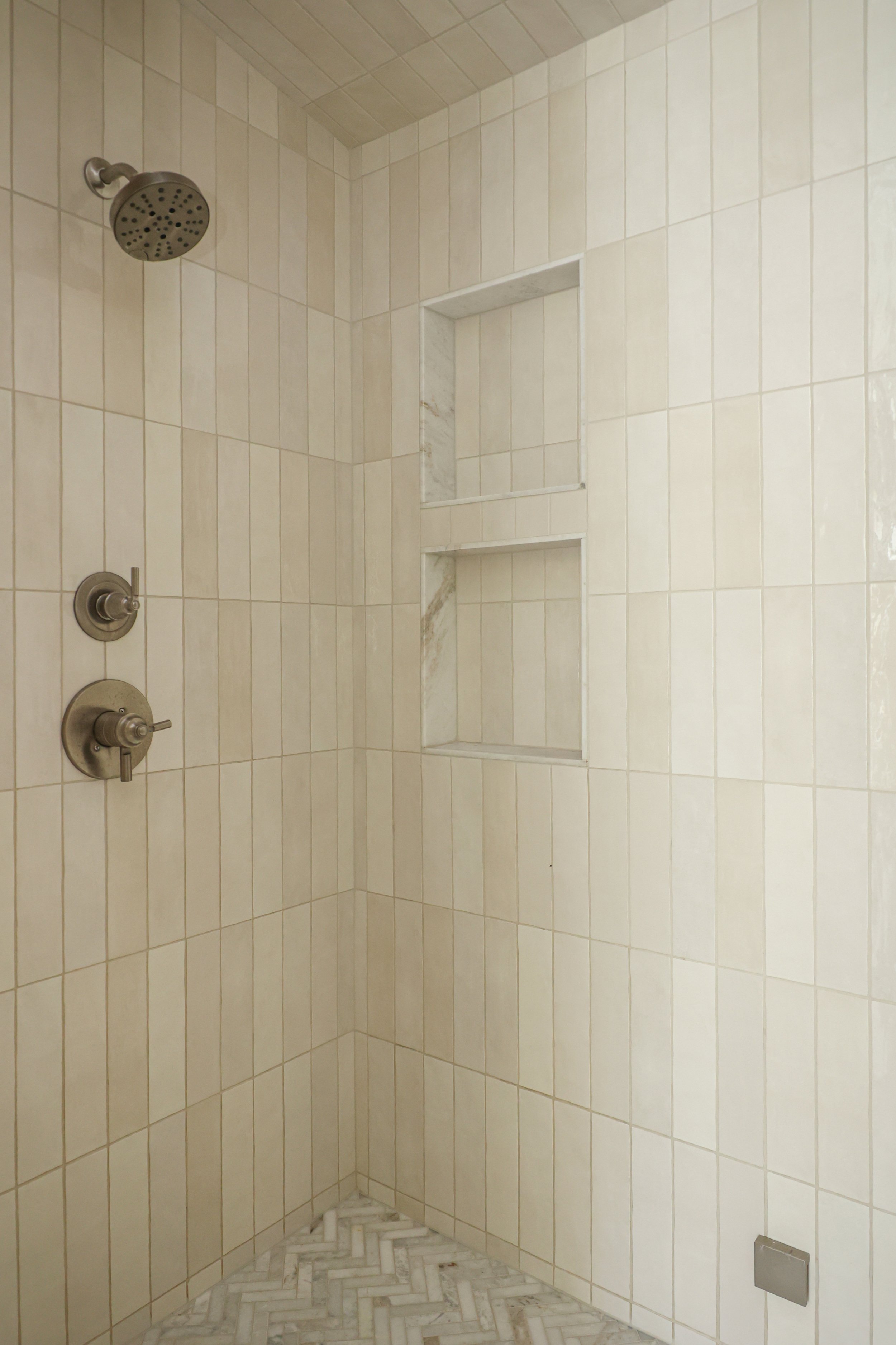 A walk-in shower with beige tiled walls, a built-in niche, a stainless steel showerhead, and a marble-patterned floor.