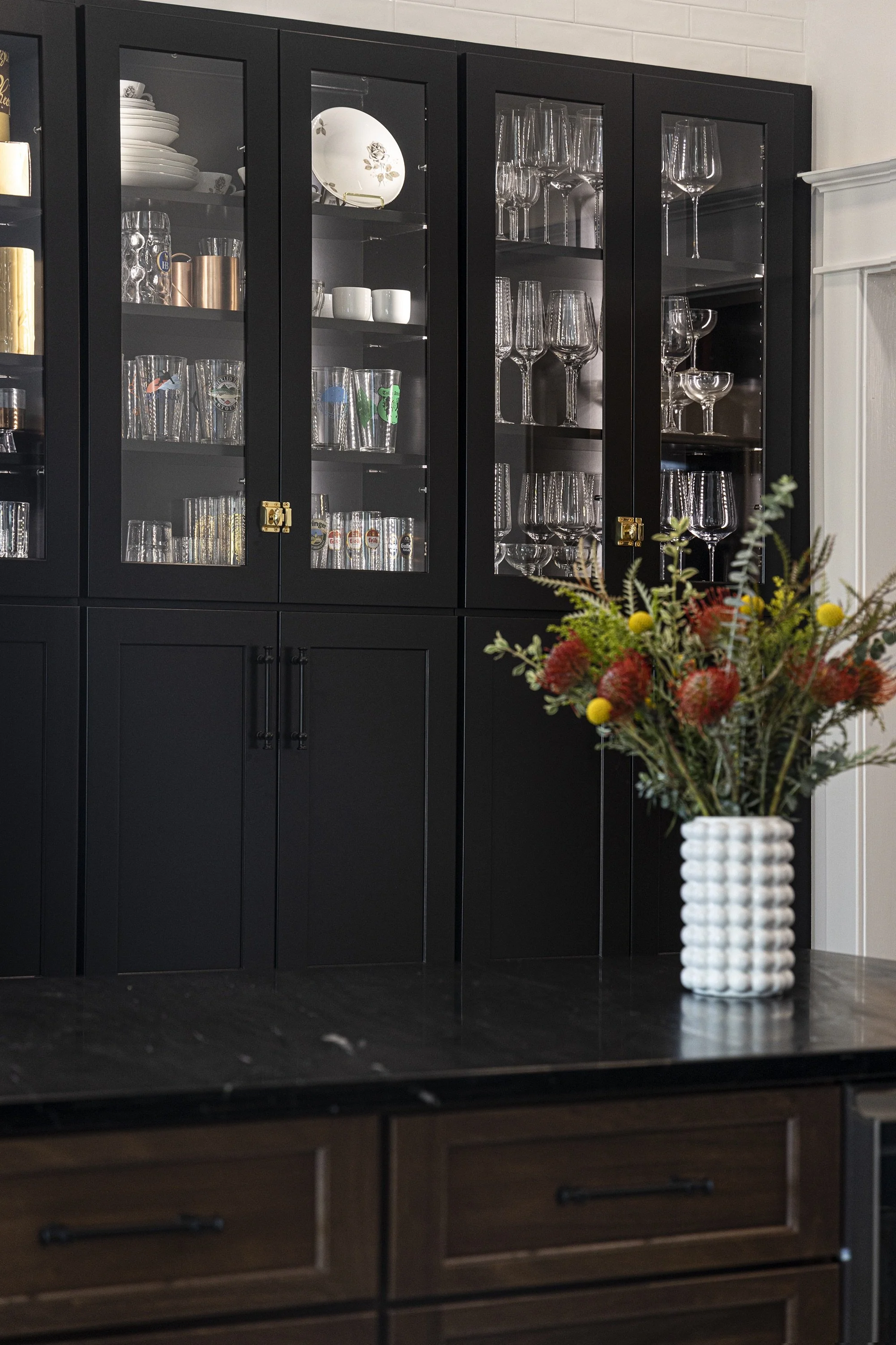A black kitchen cabinet with glass doors displaying glassware and dishes, with a white vase containing a colorful flower arrangement on a dark countertop in front.