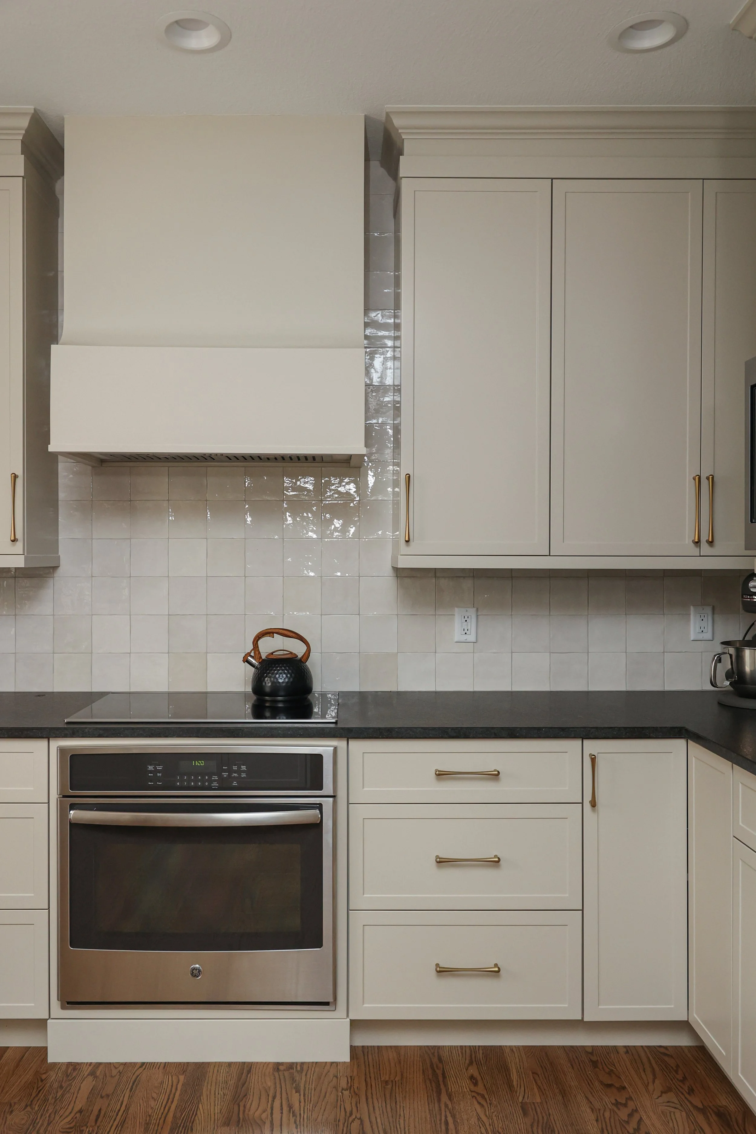 Modern kitchen with cream-colored cabinets, black countertop, stainless steel oven, black kettle on stove, beige tiled backsplash, and hardwood floor.