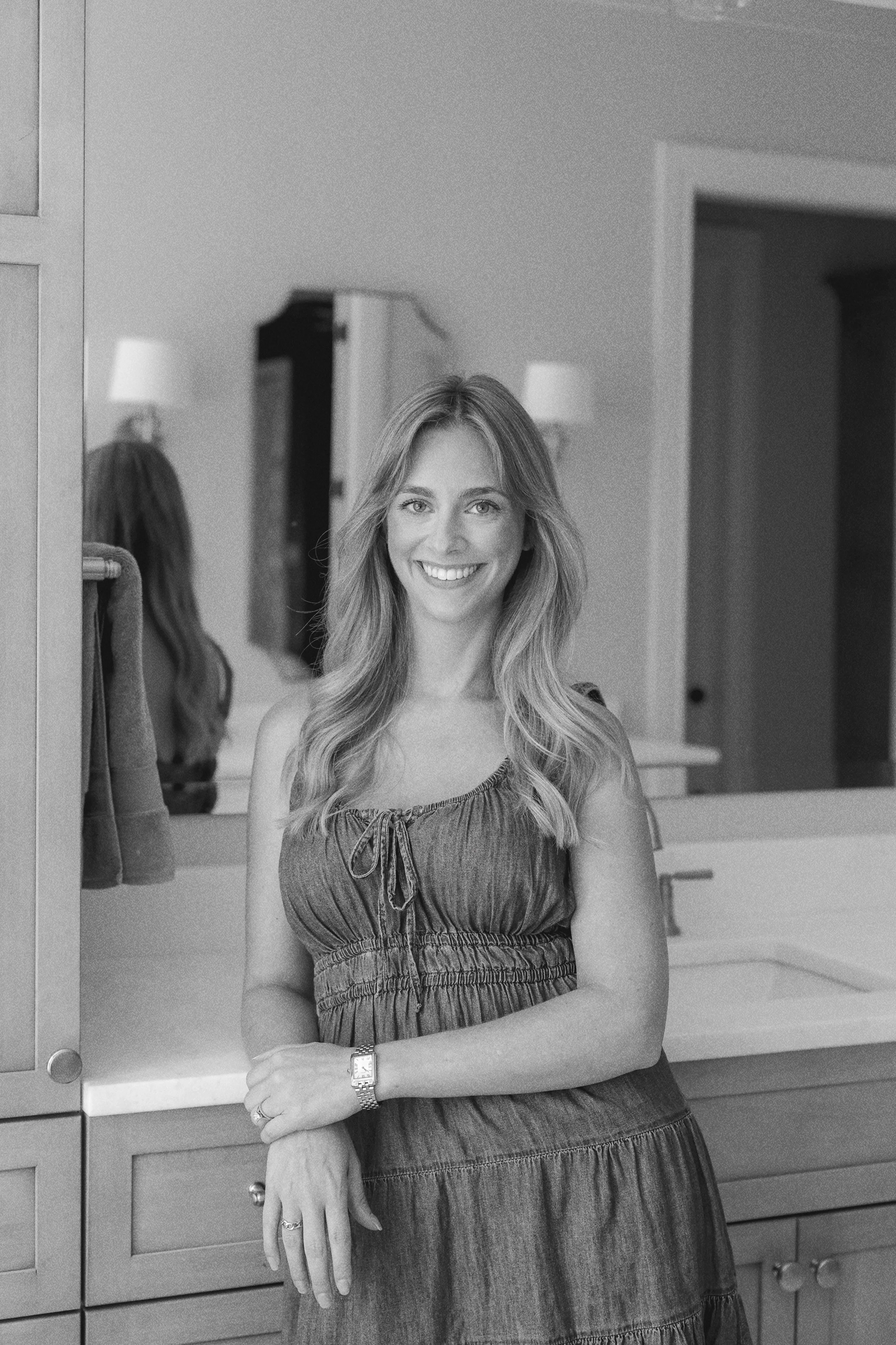 A woman with long wavy hair smiling and standing in a bathroom with a mirror behind her reflecting her back, on a countertop, and open cabinets.
