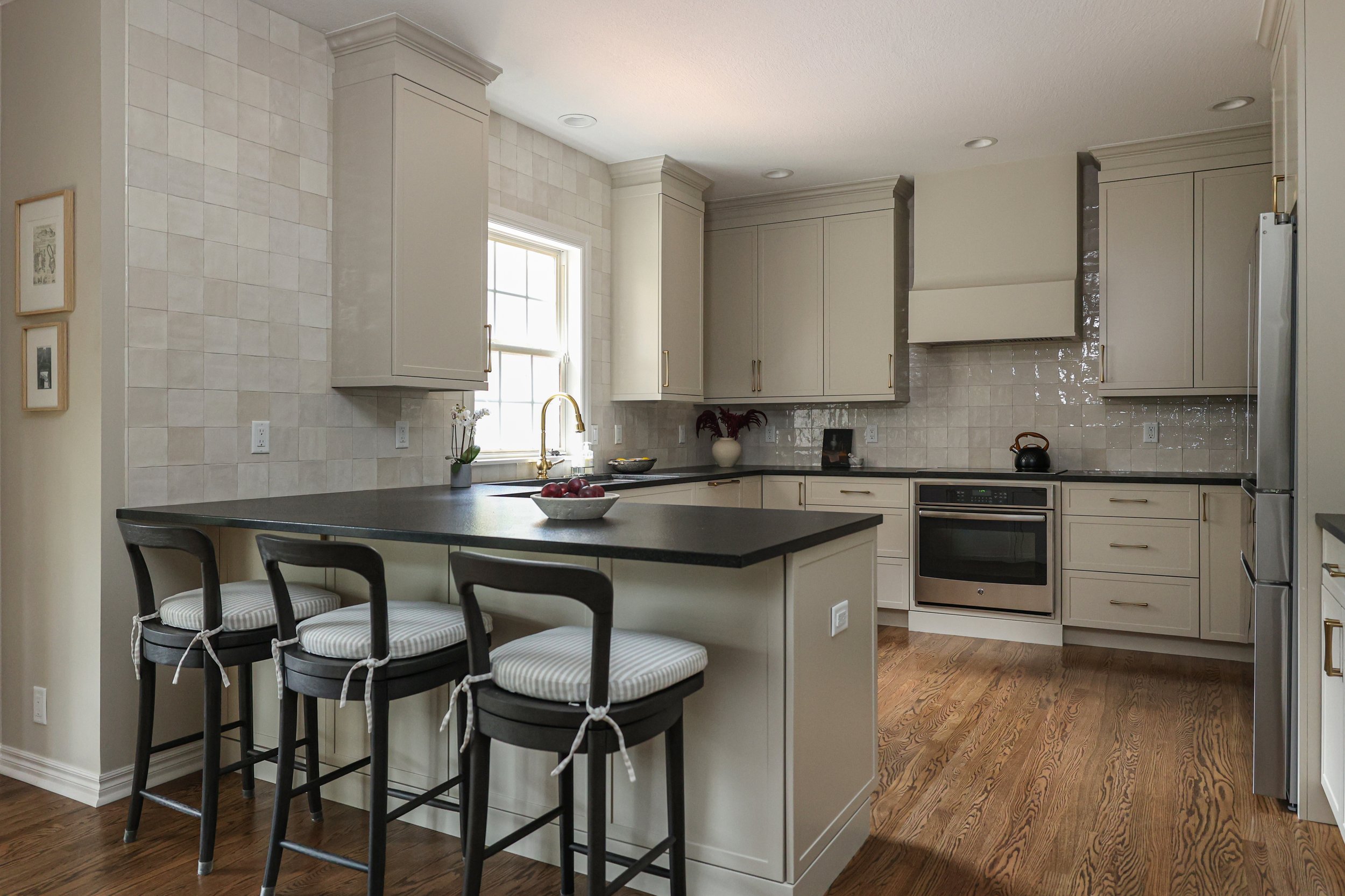 Modern kitchen with cream cabinets, black countertop island, wooden floor, and stainless steel appliances, including oven and refrigerator, with a window above the sink and three barstools with striped cushions.