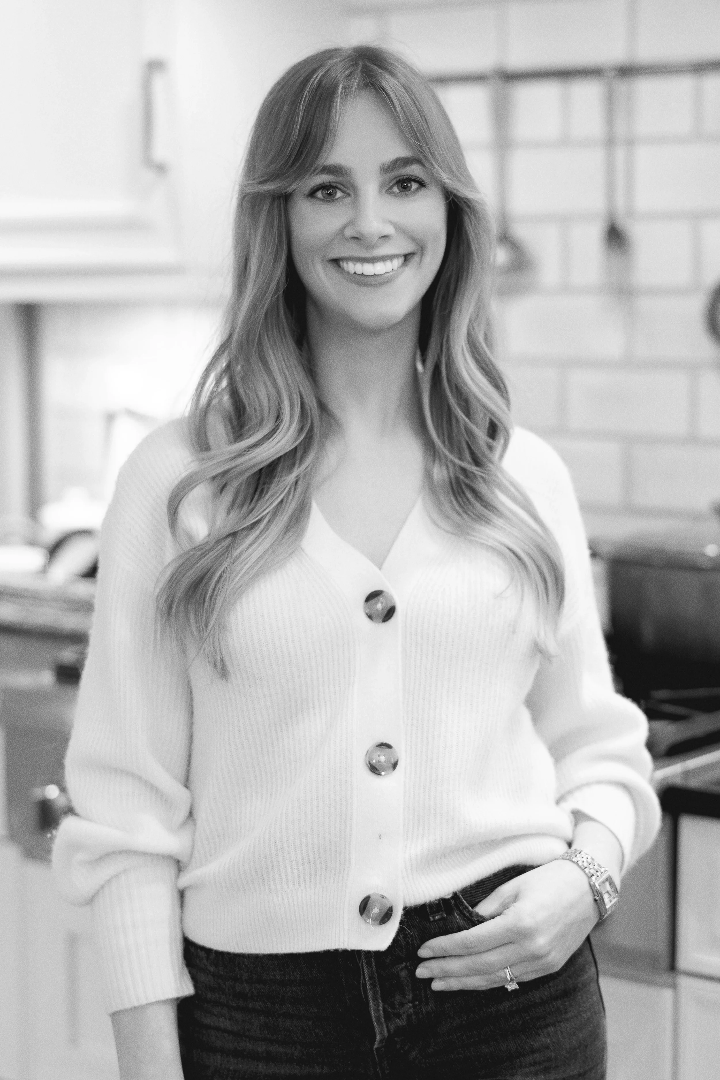 A woman with long wavy hair, smiling, wearing a white cardigan with large buttons and dark jeans, standing in a kitchen.