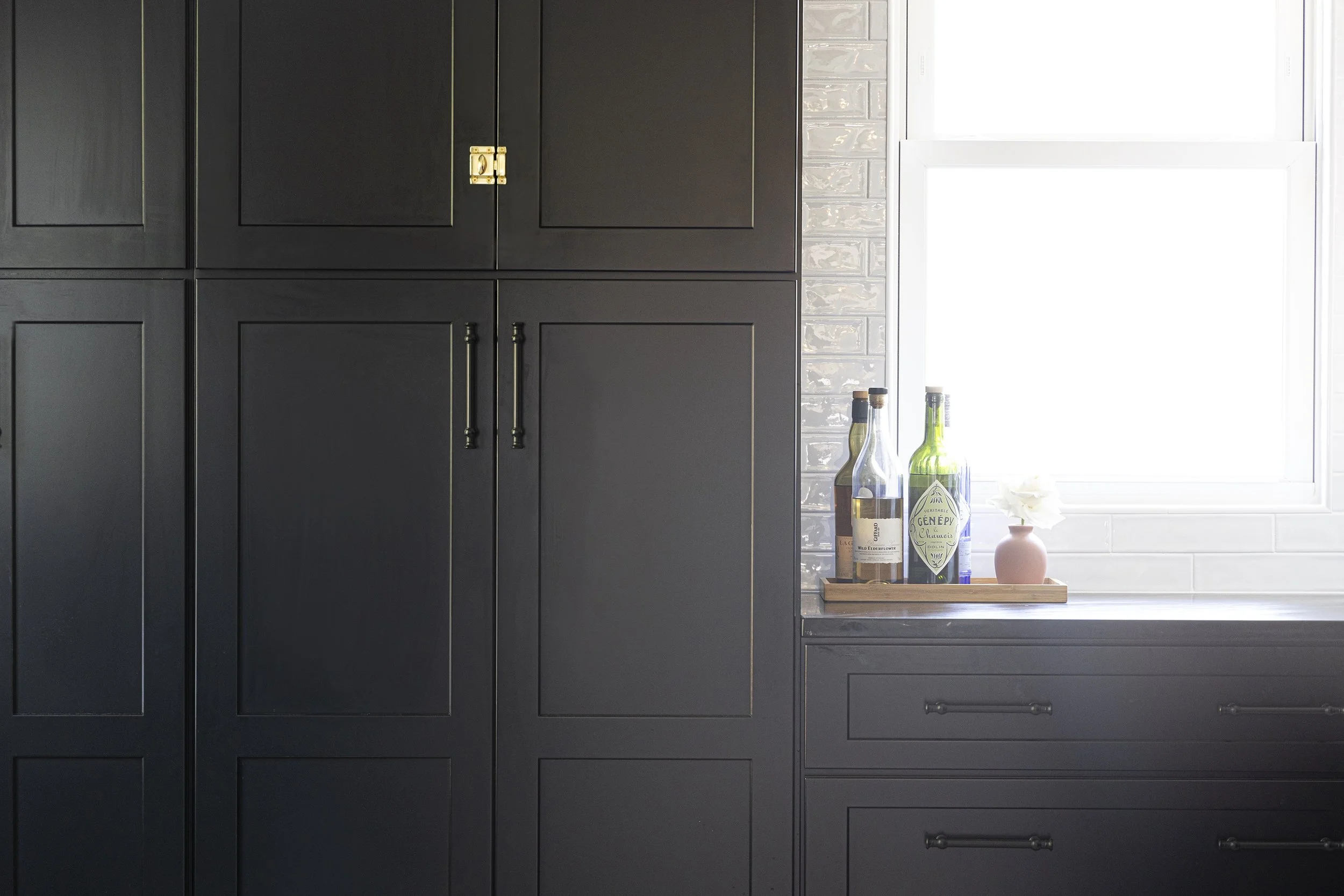 Black kitchen cabinets with handles and a window with bottles and a small vase with a flower on the windowsill.