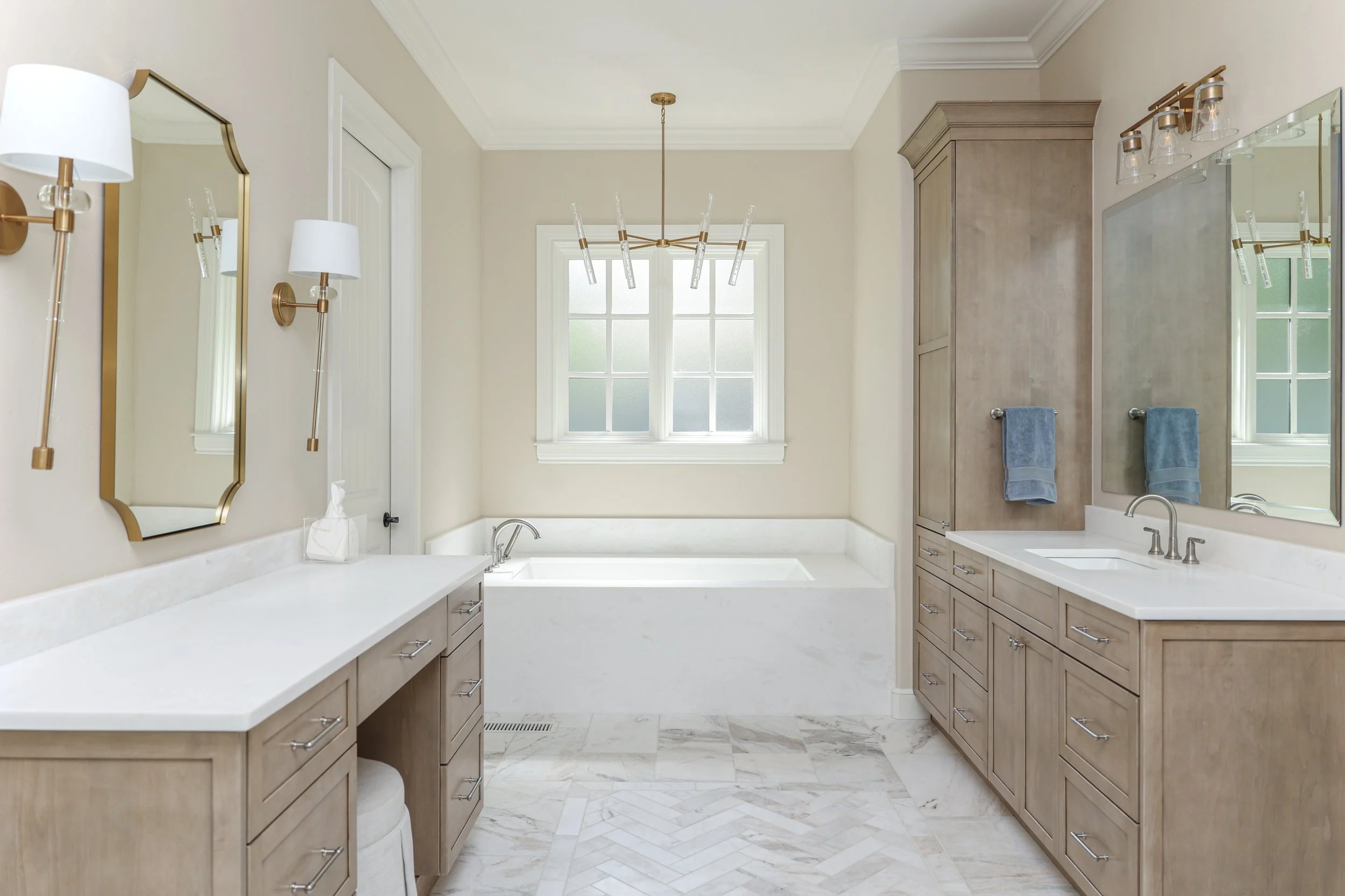 Modern bathroom with double sinks, large mirror, towel racks, a bathtub beneath a window, and beige cabinetry.
