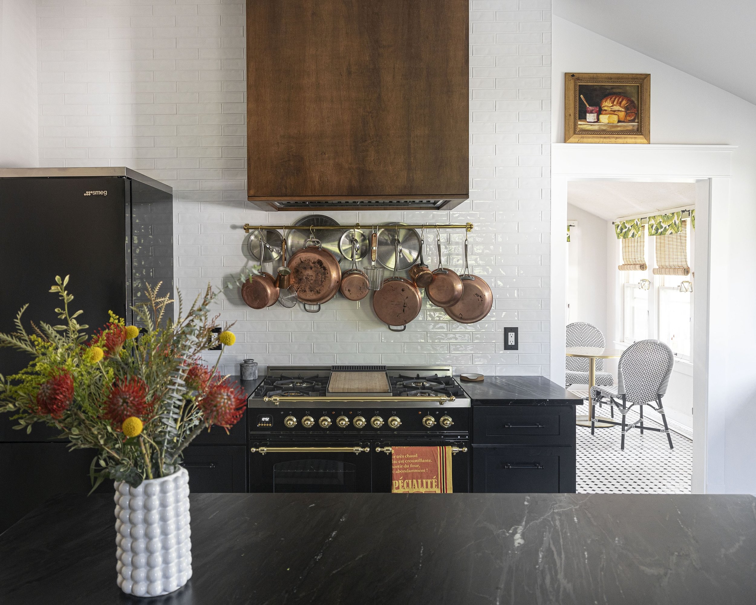 A kitchen with copper pots hanging above a black stove, white brick backsplash, dark countertop, a vase of colorful flowers on the counter, and a view into a bright dining area with a patterned table and chairs.