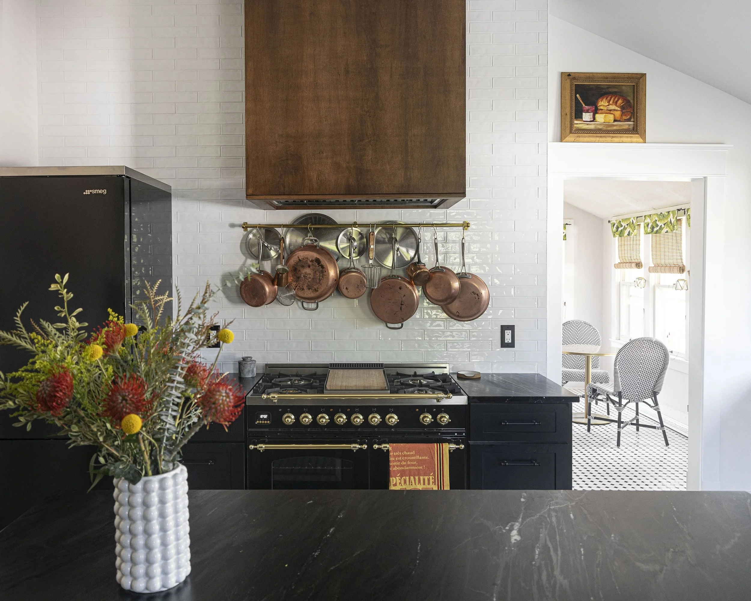 A kitchen with a black stove, copper pots hanging on a rail above it, a bouquet of flowers in a white vase on the black countertop, and a view into a bright dining area with a patterned floor and two chairs next to windows.