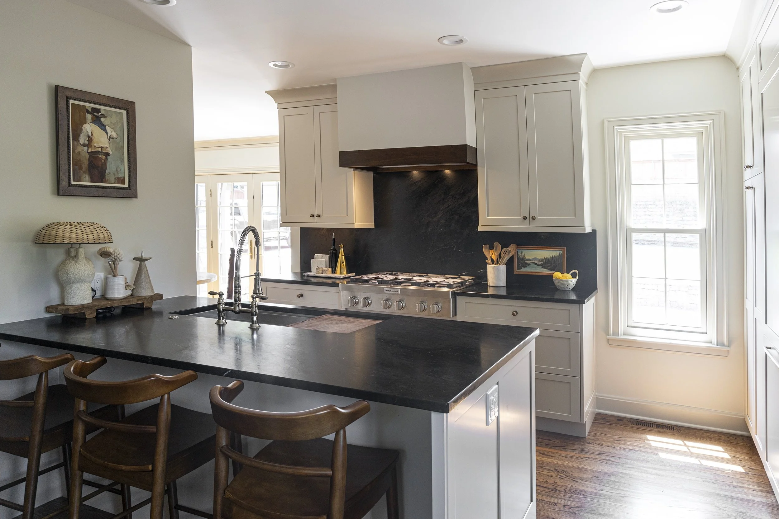 Modern kitchen with black countertop, white cabinets, a stainless steel stove, a window letting in natural light, and wooden bar stools.