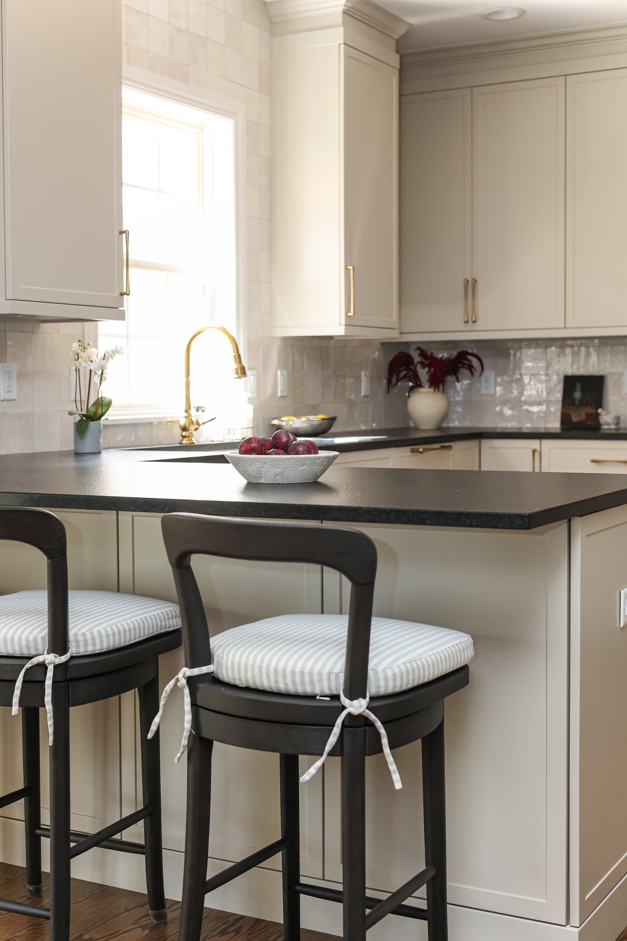 Kitchen with cream cabinets, black countertop, gold hardware, a window with sunlight, a gold faucet, a white bowl with red apples, and bar stools with striped cushions.