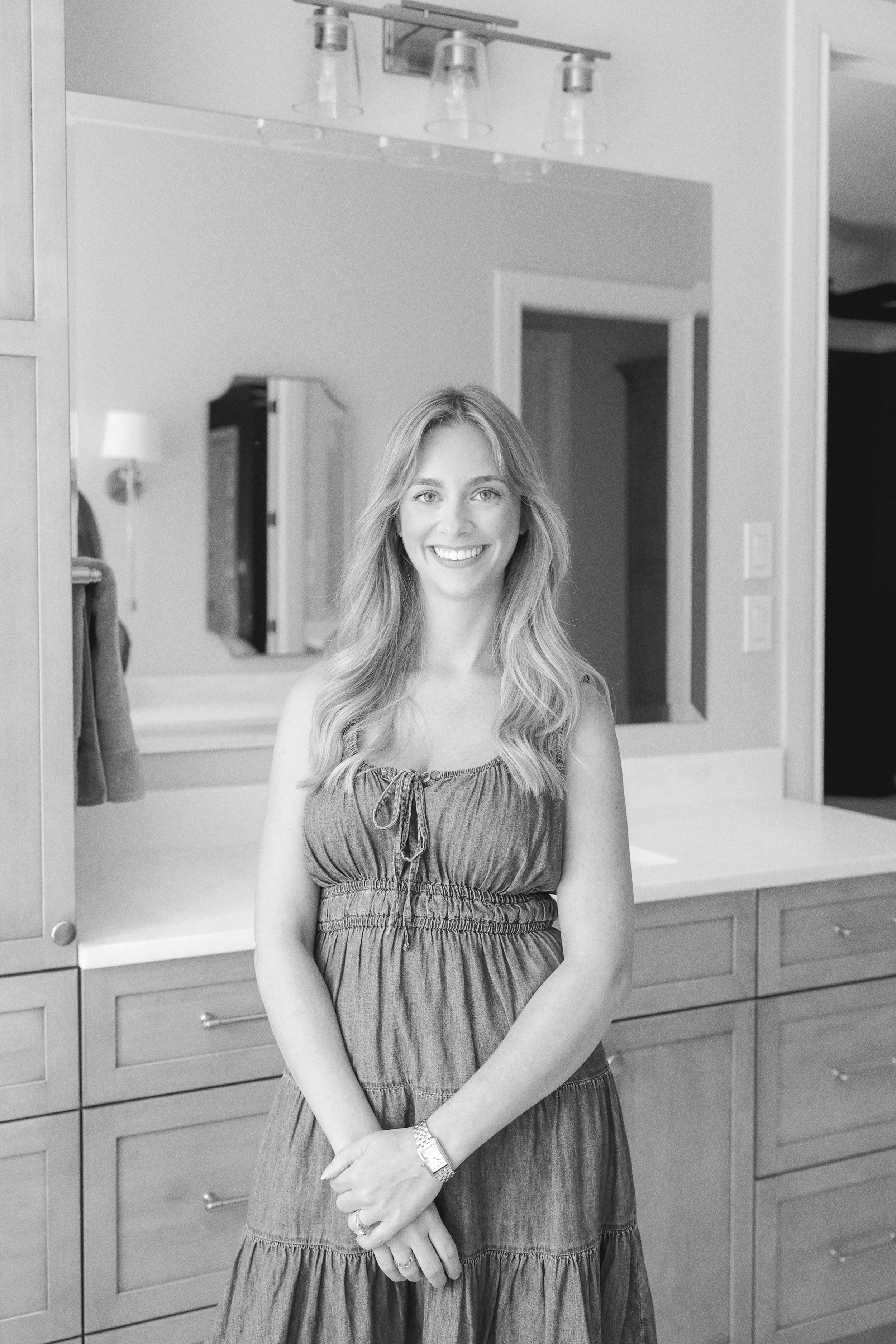 A woman with long wavy hair smiling and standing indoors in front of a wooden dresser with drawers, in a room with a mirror, a wall with switches, and a door.