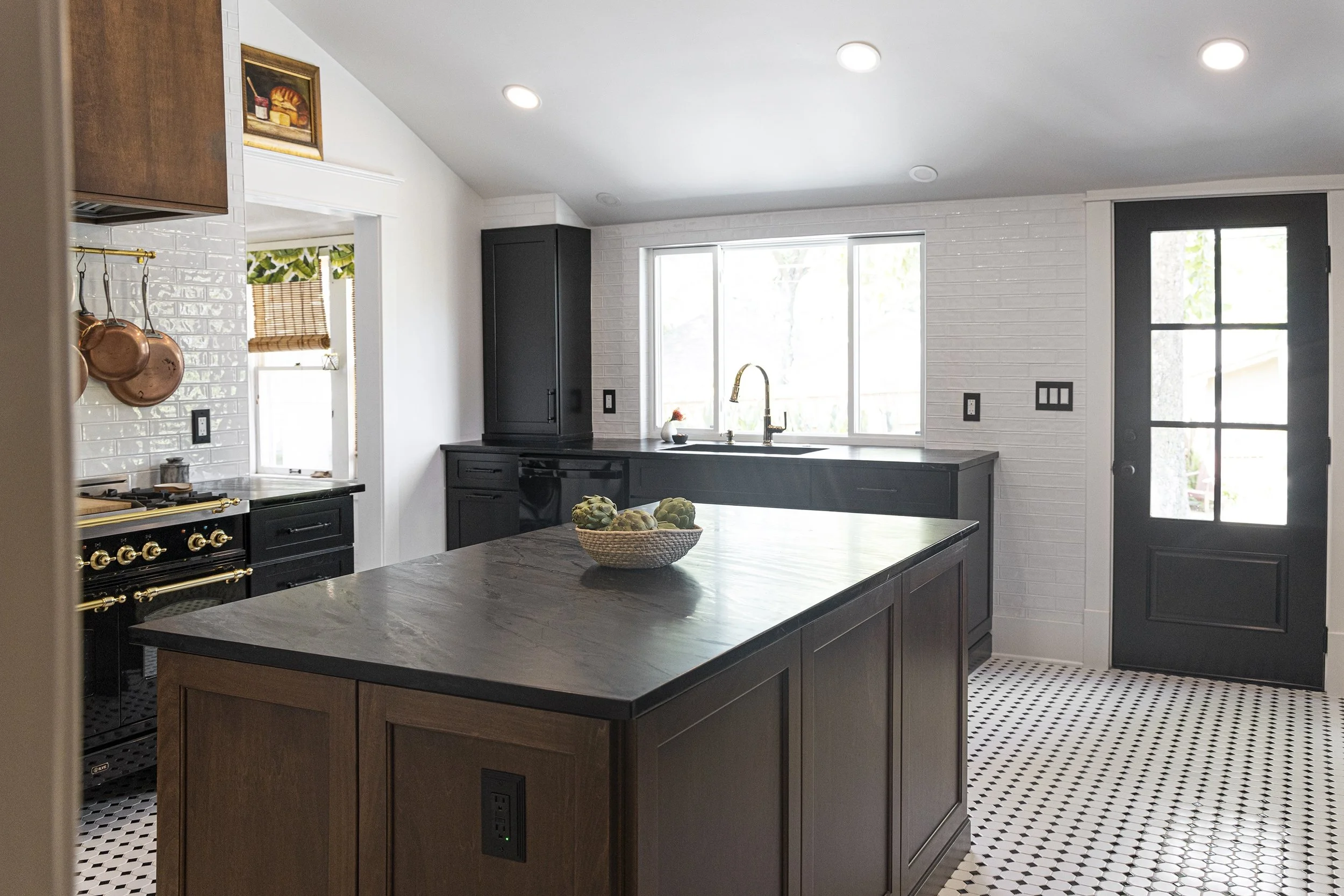 Modern kitchen with black cabinets, a wooden kitchen island, white subway tile backsplash, copper pots hanging, a window above the sink, and a black door with multiple glass panes.