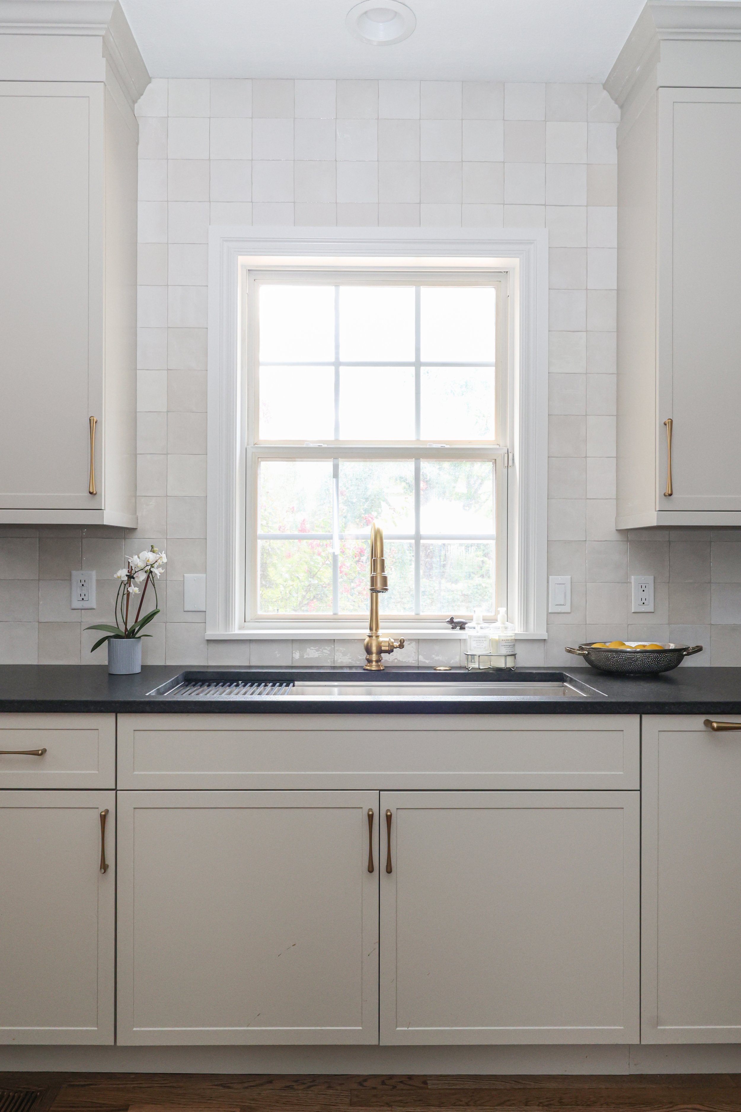A kitchen sink area with a window above, a gold faucet, cream cabinets with gold handles, a small potted orchid on the left, and a bowl of lemons on the right.
