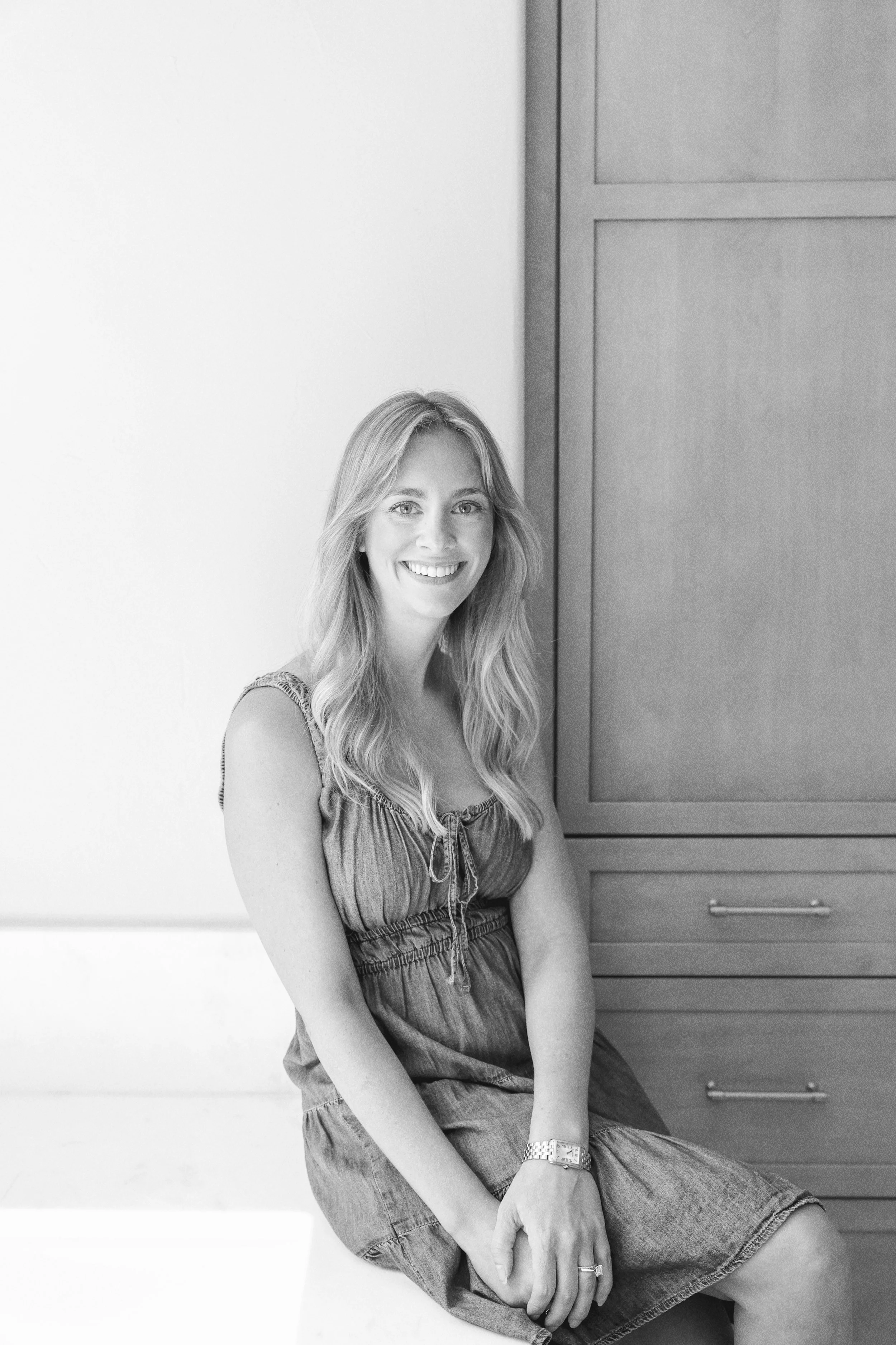 A smiling woman with long wavy hair sitting on a white surface, wearing a sleeveless dress, with a wooden cabinet in the background.