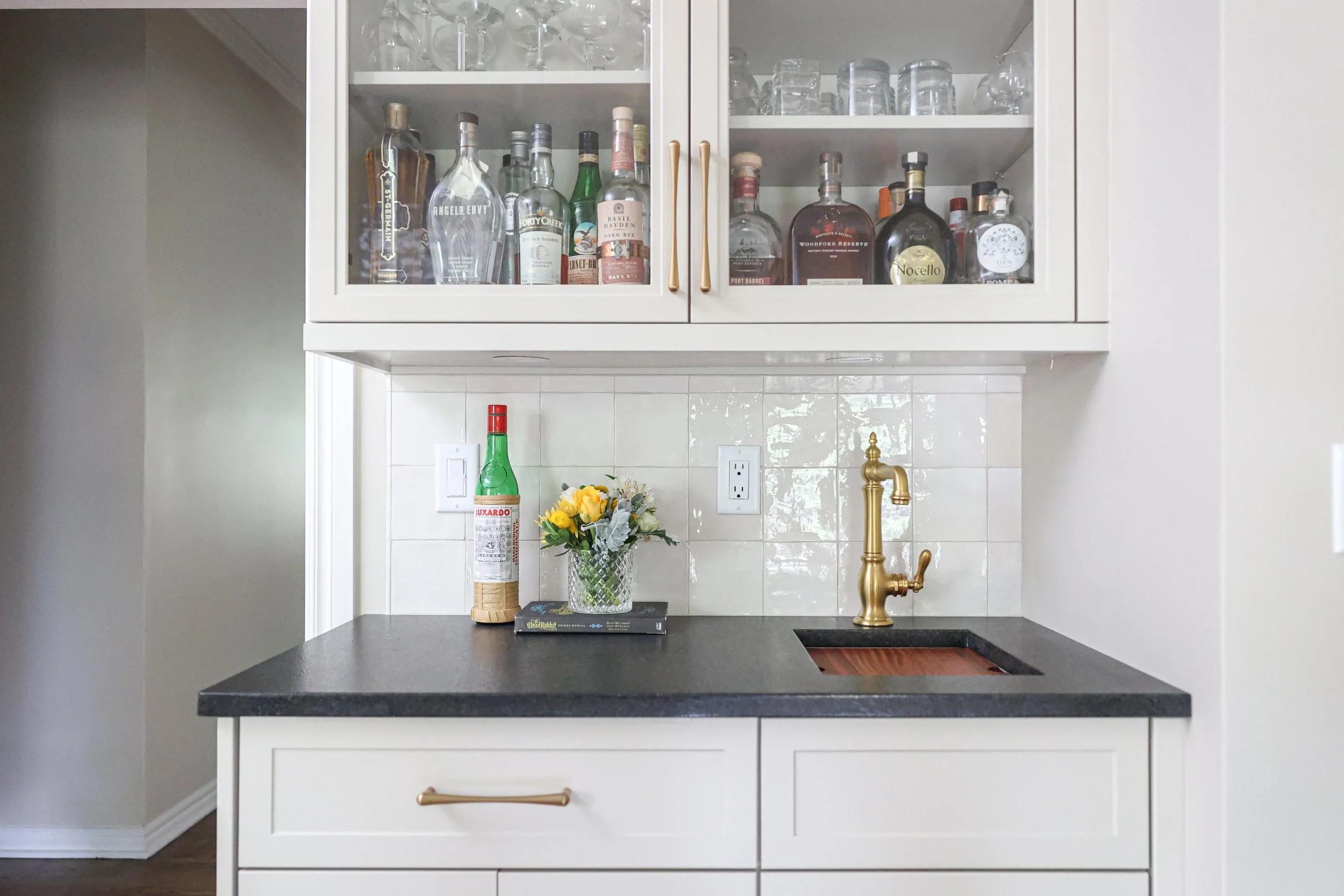 Kitchen bar area with a countertop, a gold faucet, a small flower arrangement, a bottle of hot sauce, and a book, with glassware and bottles of alcohol in a glass-front cabinet above.