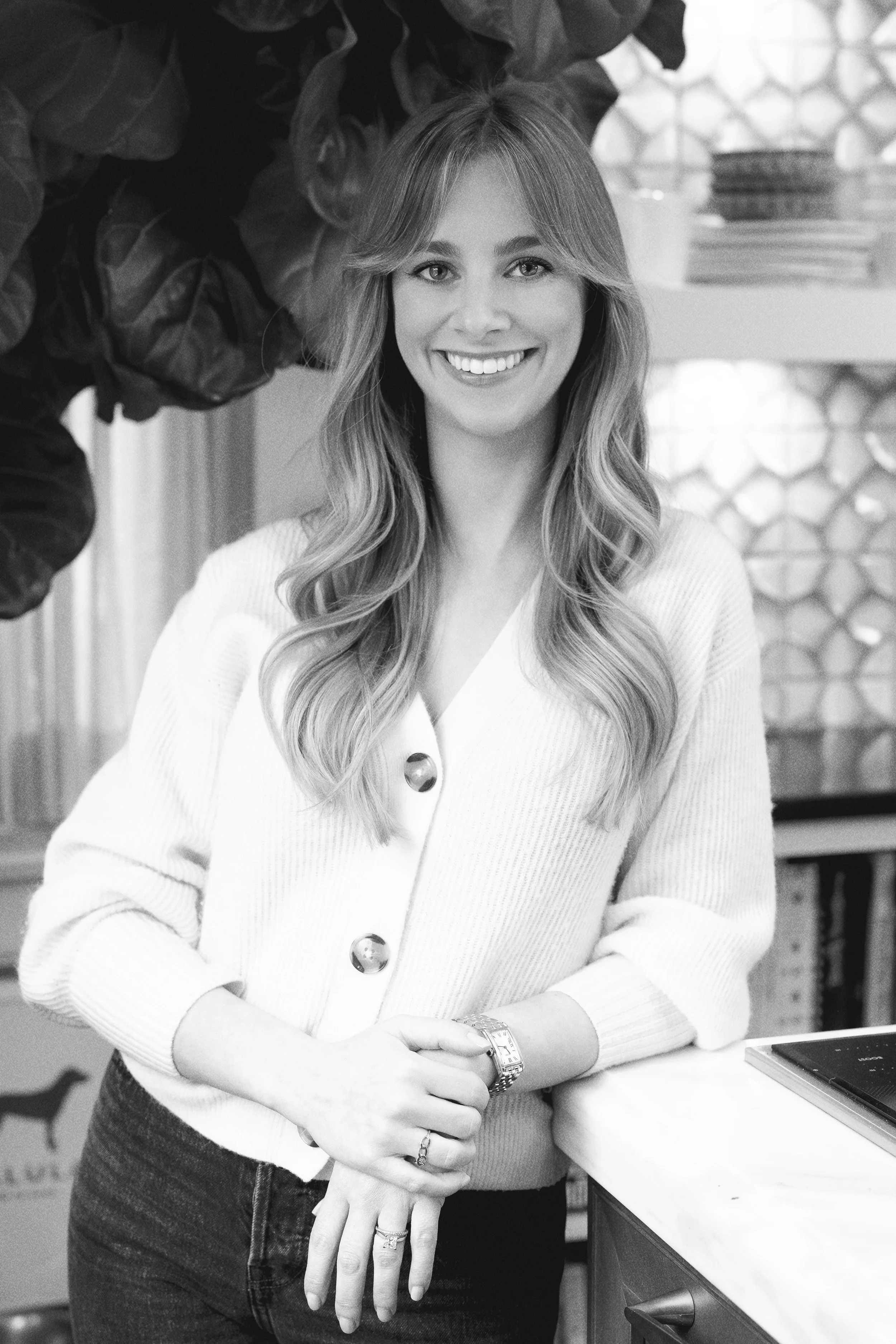 A woman with wavy hair, smiling, wearing a light-colored cardigan with large buttons, standing with her arms folded on a counter, in a room with indoor plants and a patterned glass window in the background.
