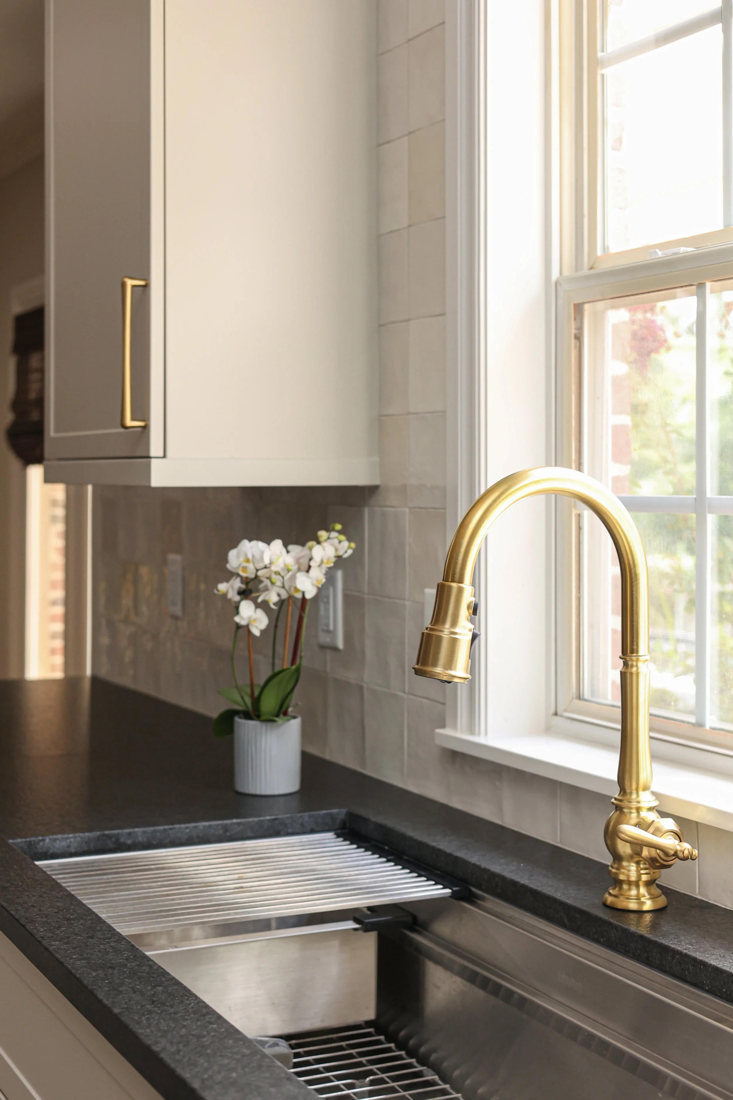 A kitchen sink with a modern gold faucet, a potted white orchid on the black countertop, and a window with sunlight streaming in.
