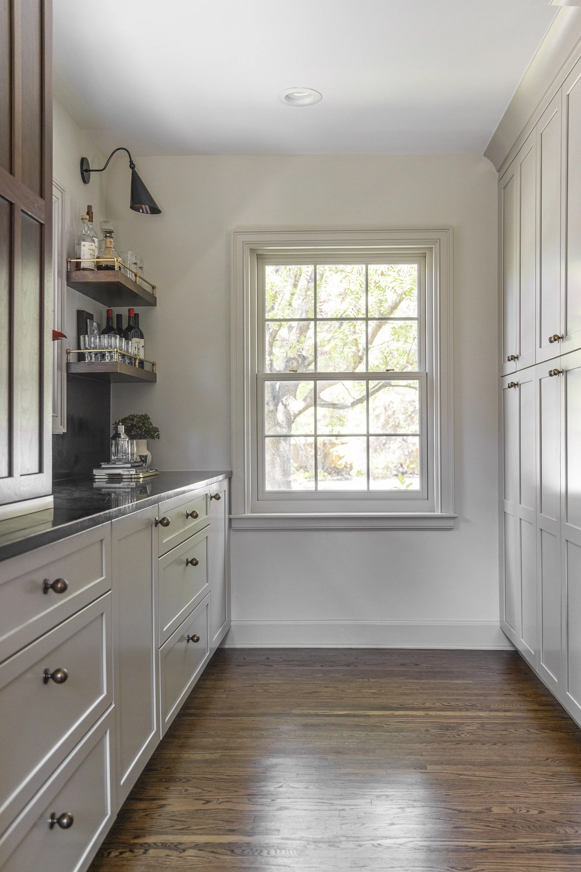 A bright kitchen with white cabinets, a large window, wooden floors, and open shelves holding bottles, glasses, and decorative items.