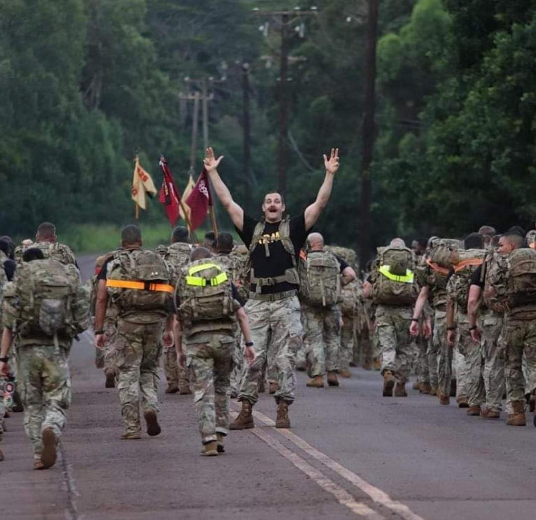 A man standing in front of a group of soldiers on a road, raising his arms in celebration or greeting, with flags behind him and trees in the background.