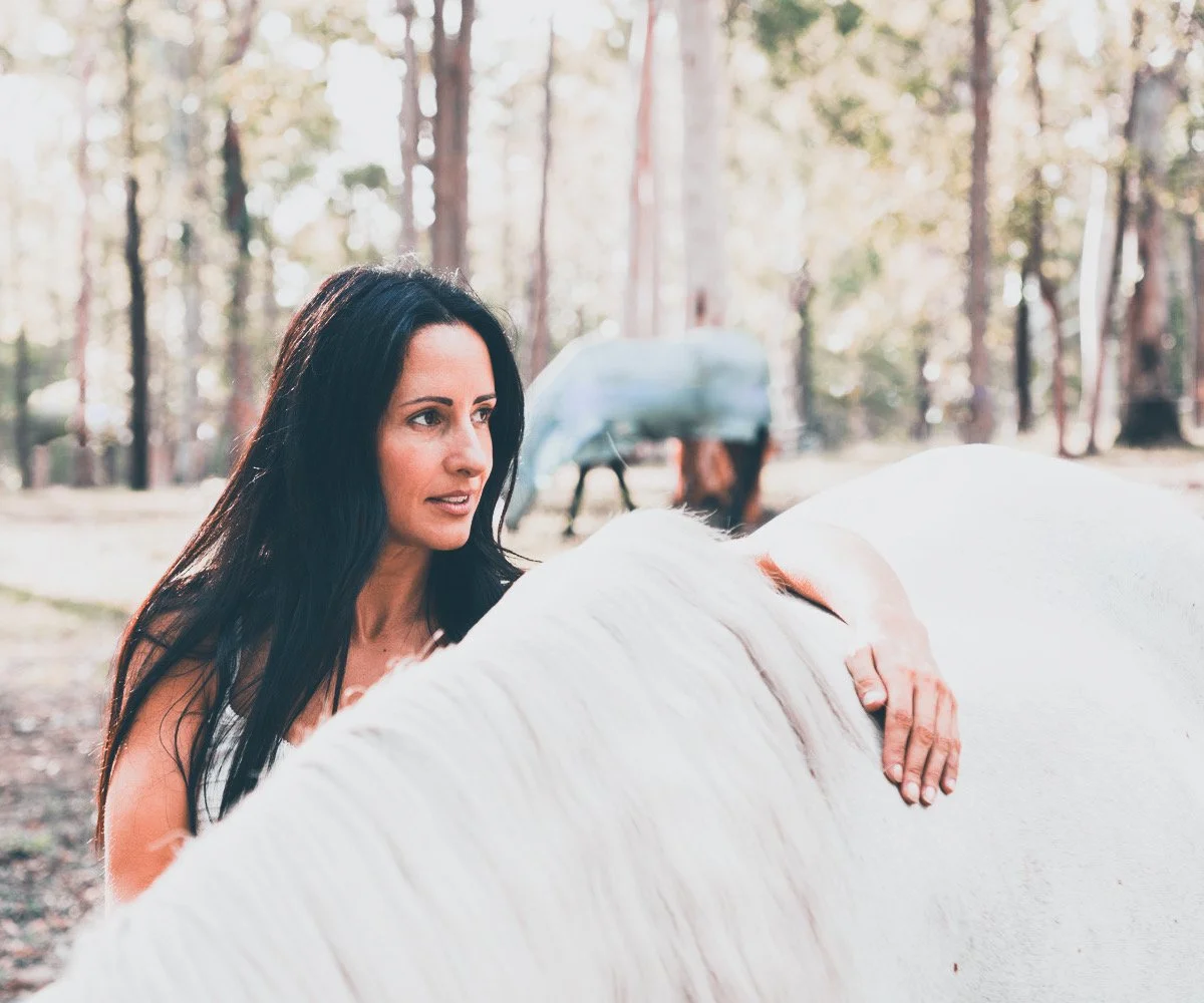 A woman with dark hair petting a white horse in a forested area.