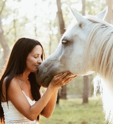 A woman with long dark hair gently holds a white horse's face, touching its nose with her hands, in a wooded outdoor setting.