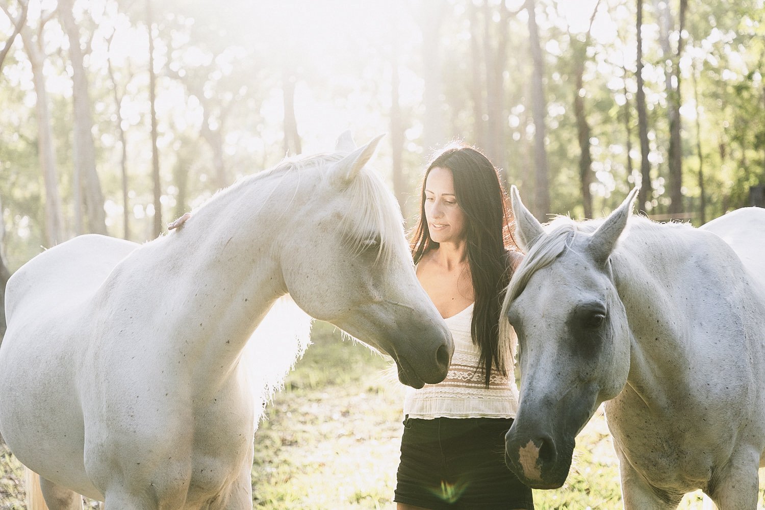 A woman with long dark hair standing outdoors in a forested area, gently touching two white horses on either side of her in the sunlight.