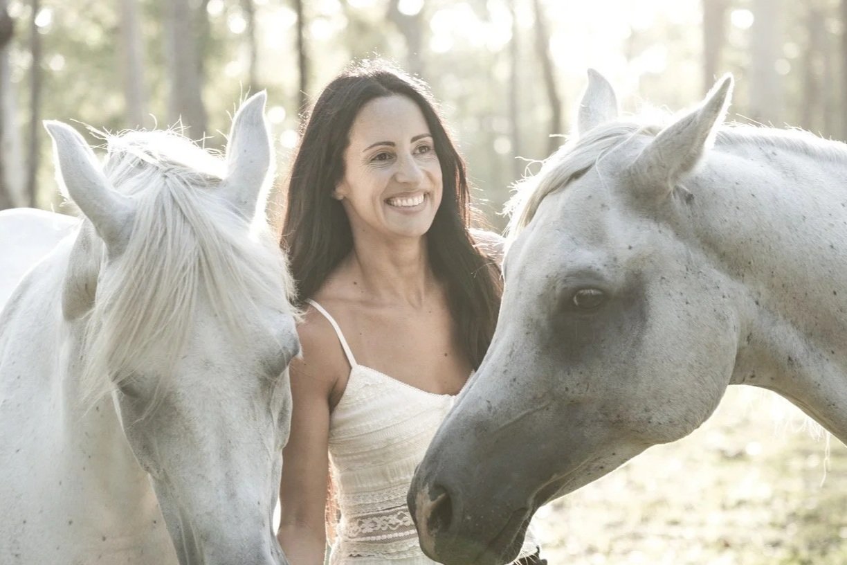 A woman smiling and standing between two white horses in a forested area.