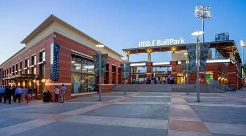 Exterior view of BB&T BallPark entrance with a few people walking in front, brick buildings, and a large gateway sign.