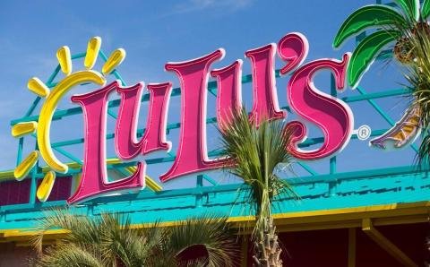 Sign reading "Lulu's" with a sun illustration in the upper left corner and palm trees in front, set against a bright blue sky.