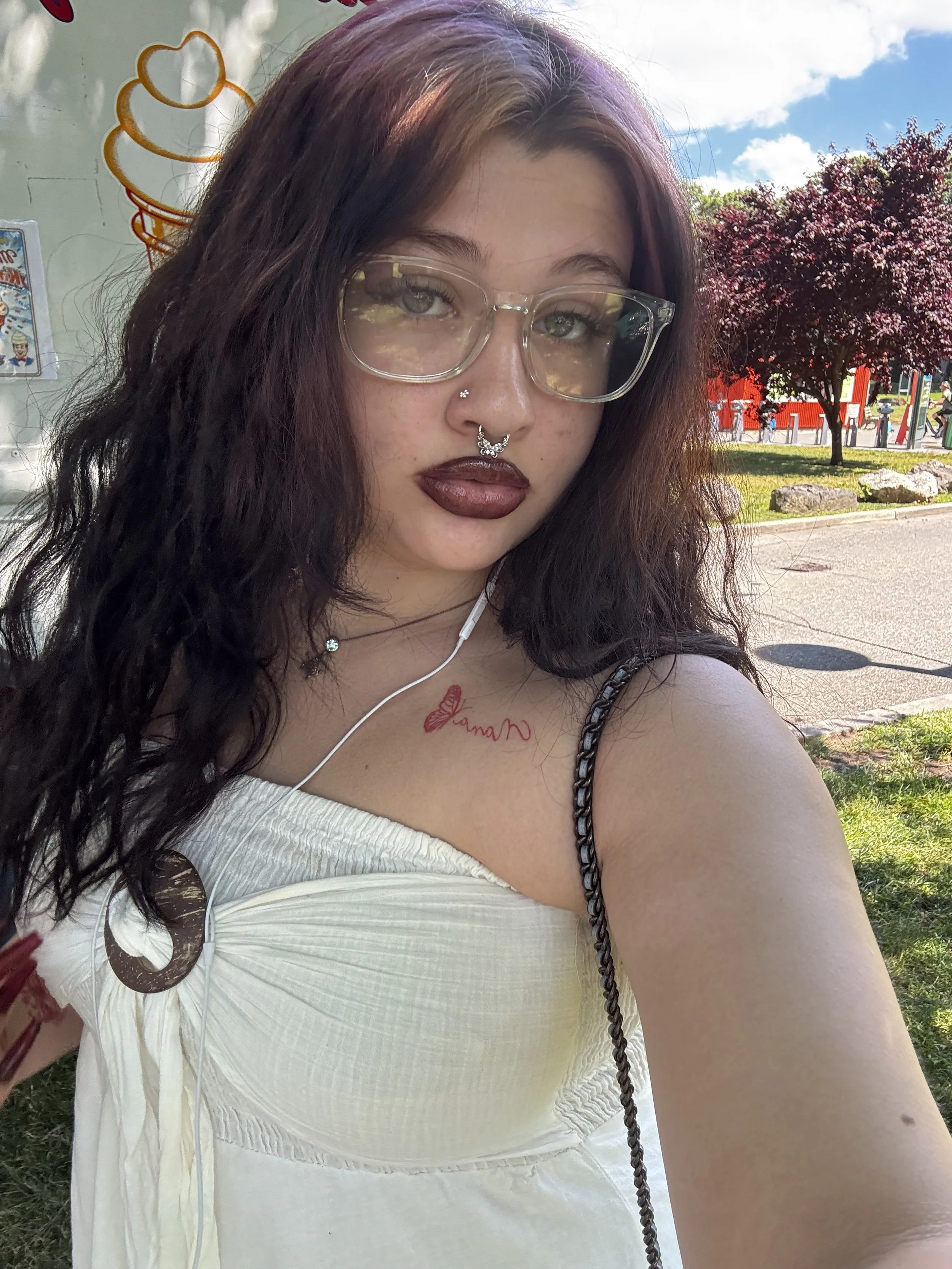 A young woman with curly dark hair, wearing clear glasses, dark lipstick, a nose ring, and a septum piercing, taking a selfie outdoors on a sunny day with a blue sky, trees, and a red fence in the background.