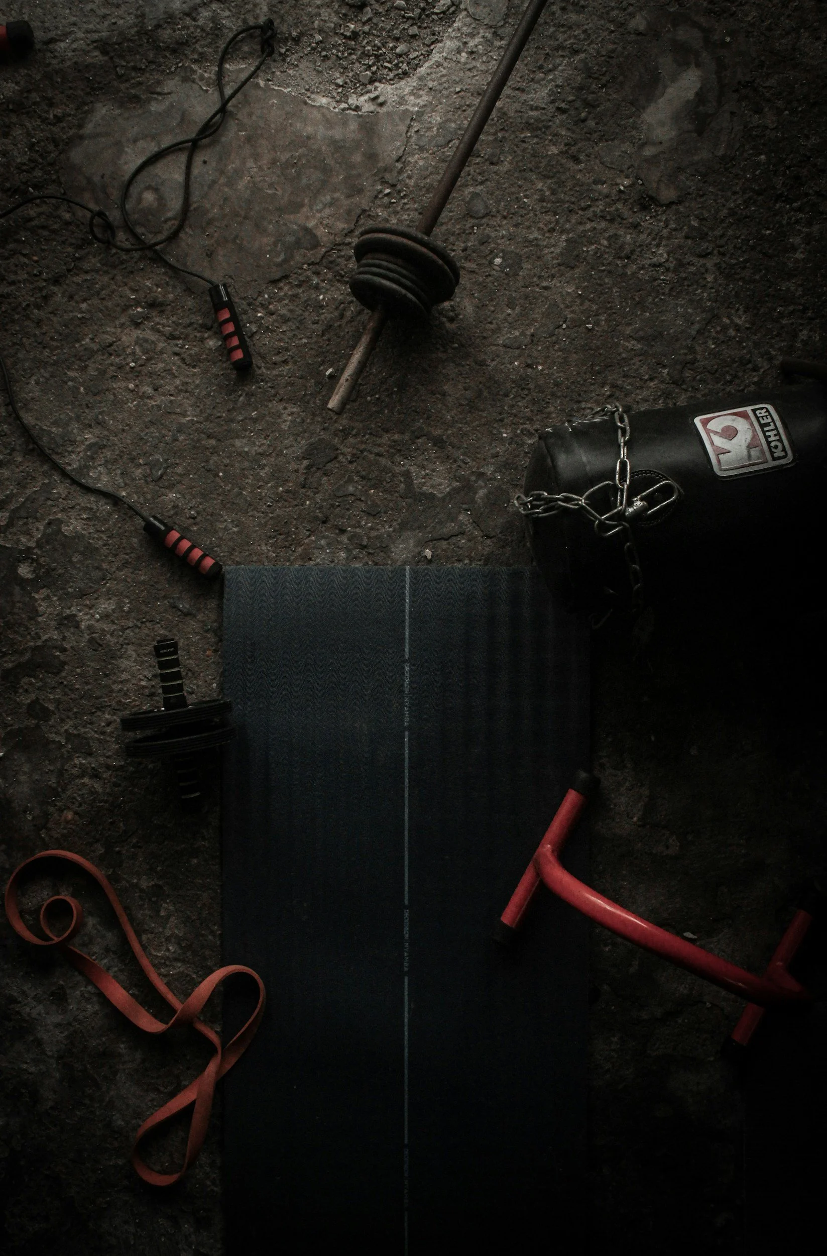 Gym equipment on a rough concrete floor, including a black yoga mat, red-handled resistance band, adjustable dumbbell, black punching bag with chain, and a barbell with weight plates.
