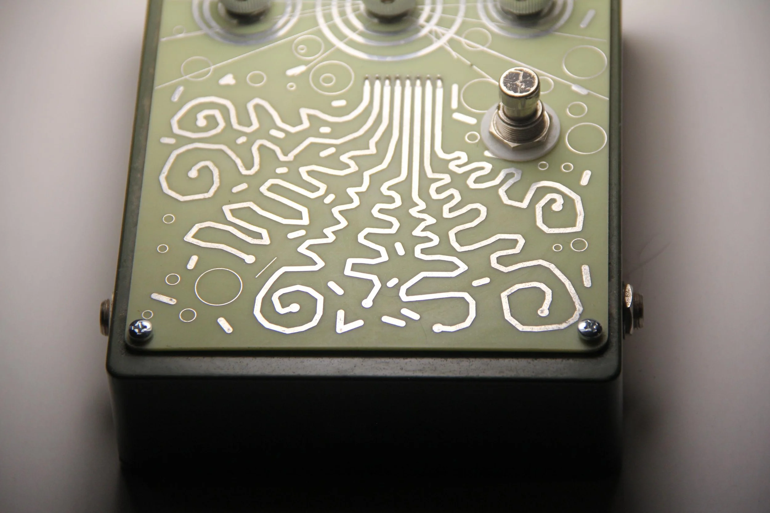 Close-up of a rectangular electronic device with a green circuit board featuring intricate white circuit patterns. There is a metal screw or bolt on the top right and some small screws at the corners. The device is placed on a white surface.