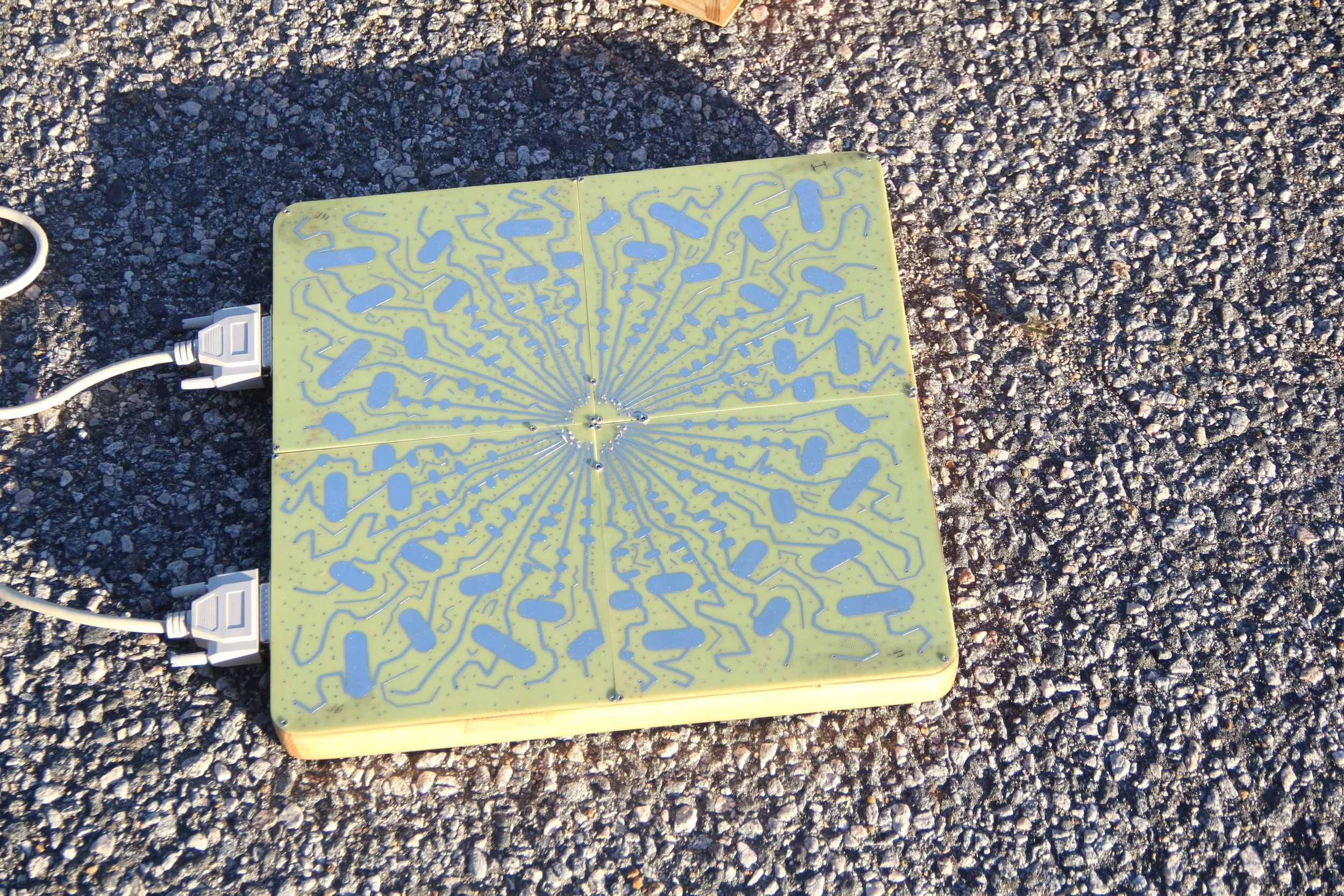 A square electronic device with a green surface and blue circuit-like pattern, connected to a power source with two white cords, placed on a gravel surface outdoors.