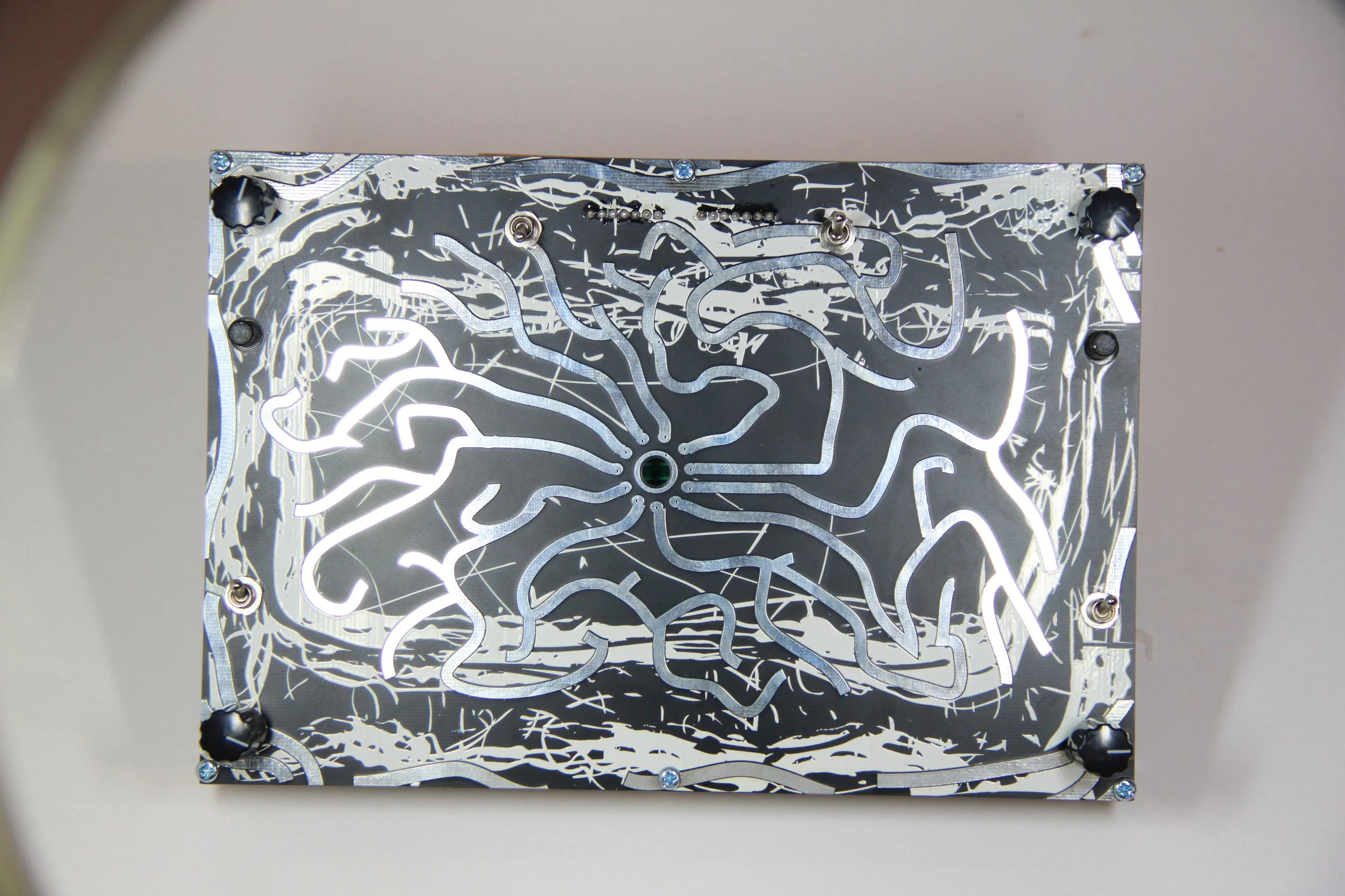A printed circuit board with various twisting and curved metallic pathways and four black securing screws at each corner.
