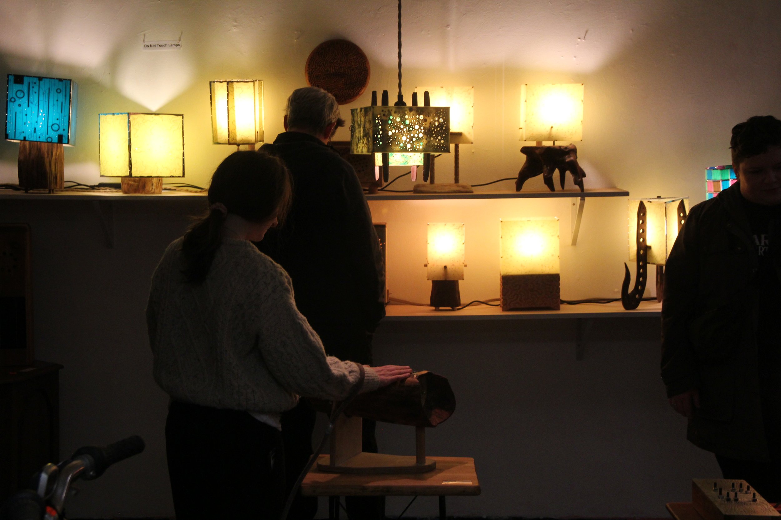 People looking at various illuminated lamps and decorative objects on display in a dimly lit room.
