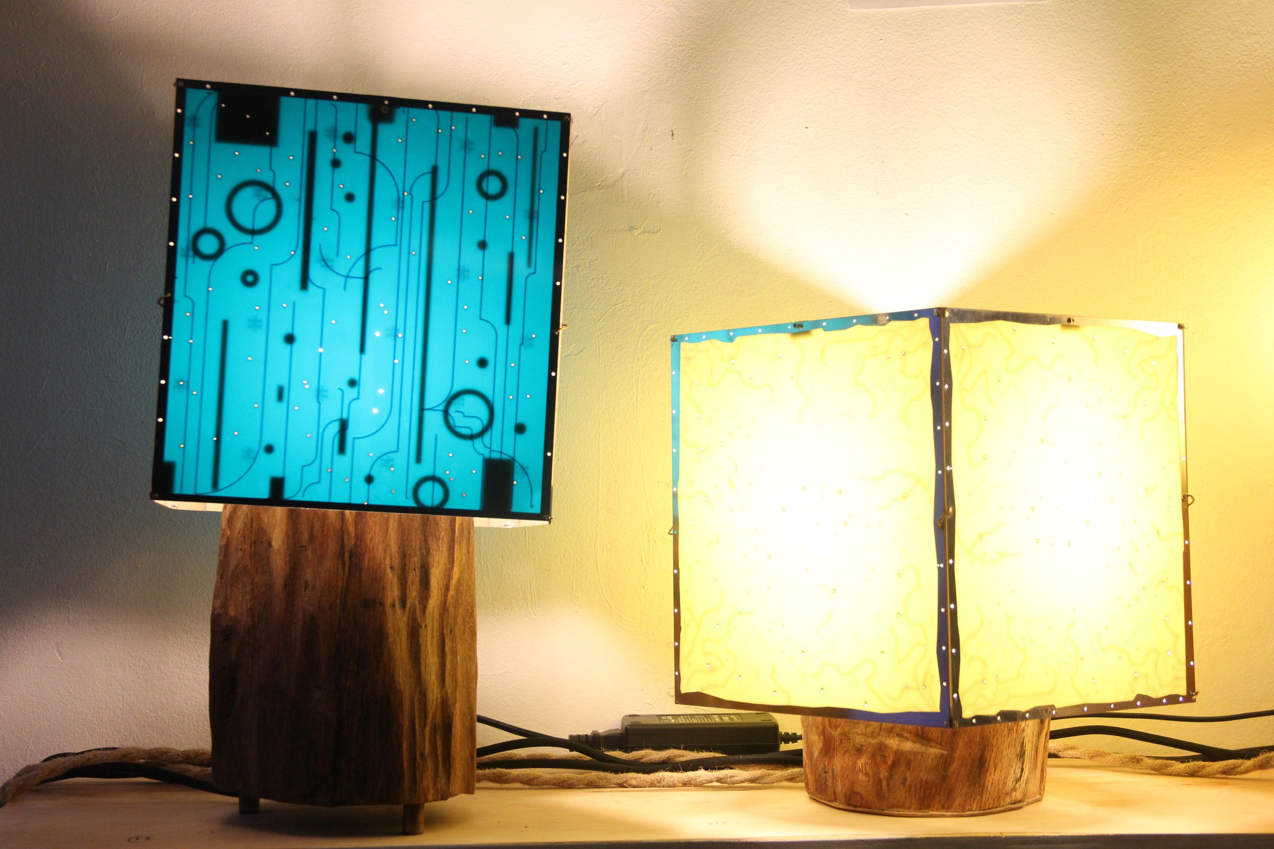 Two illuminated lamps with wooden bases, one blue with circuit-like patterns and one yellow with a textured surface, placed on a wooden surface against a wall.