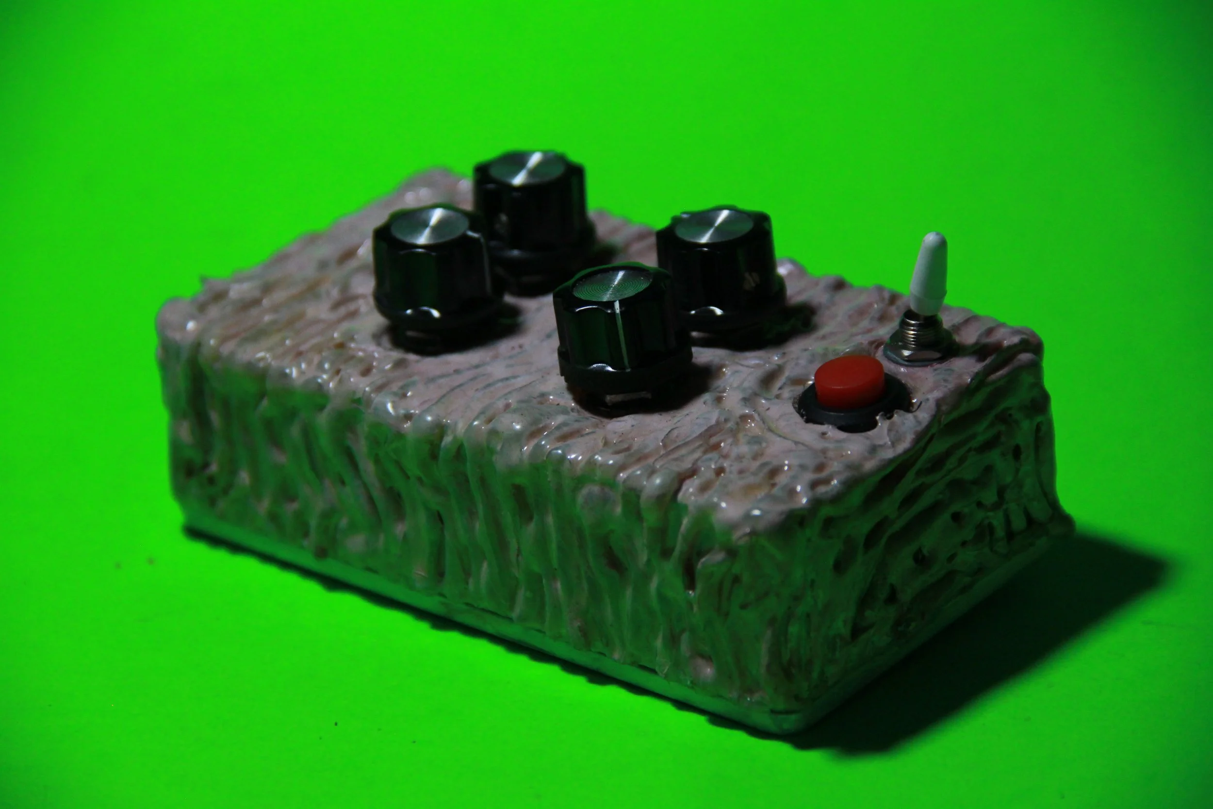 A rectangular guitar effects pedal with a textured, beige surface, black control knobs, a red button, and a white antenna against a bright green background.