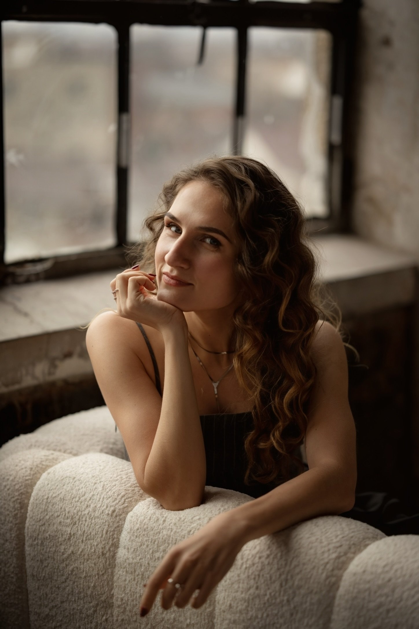 A young woman with long, wavy brown hair, resting her chin on her hand, sitting on a beige, textured sofa near a window with a blurred outdoor background.