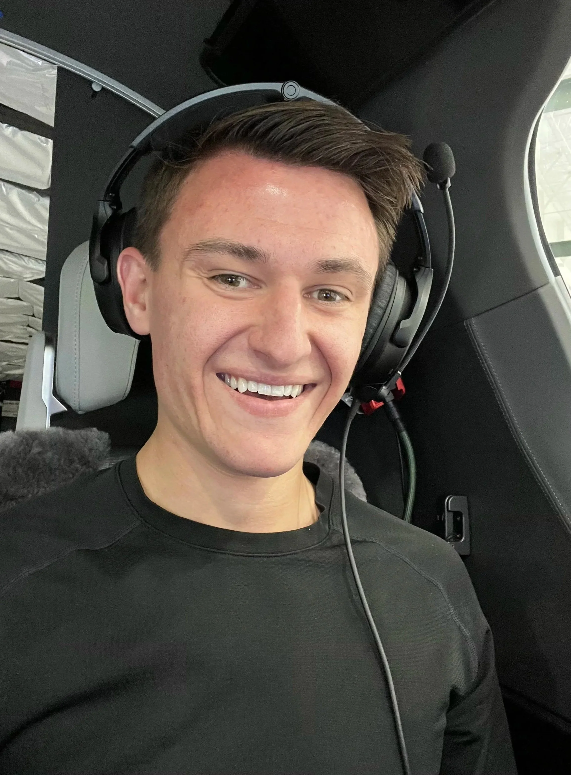 A young man sitting in an aircraft, smiling, wearing an aviation headset, and a black shirt.