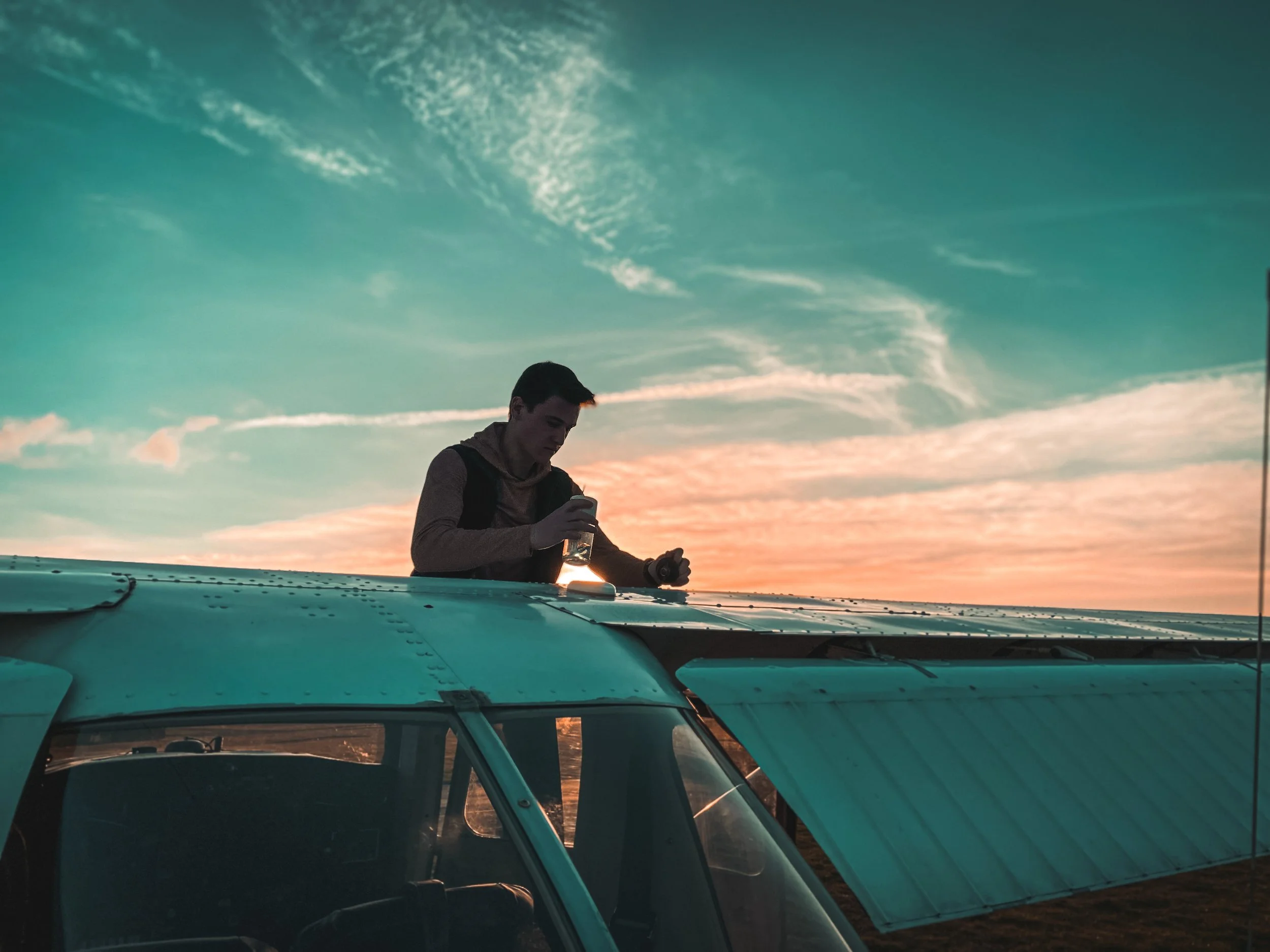A young man preflighting an aircraft holding a fuel sump, with a beautiful sunset sky with clouds in the background.