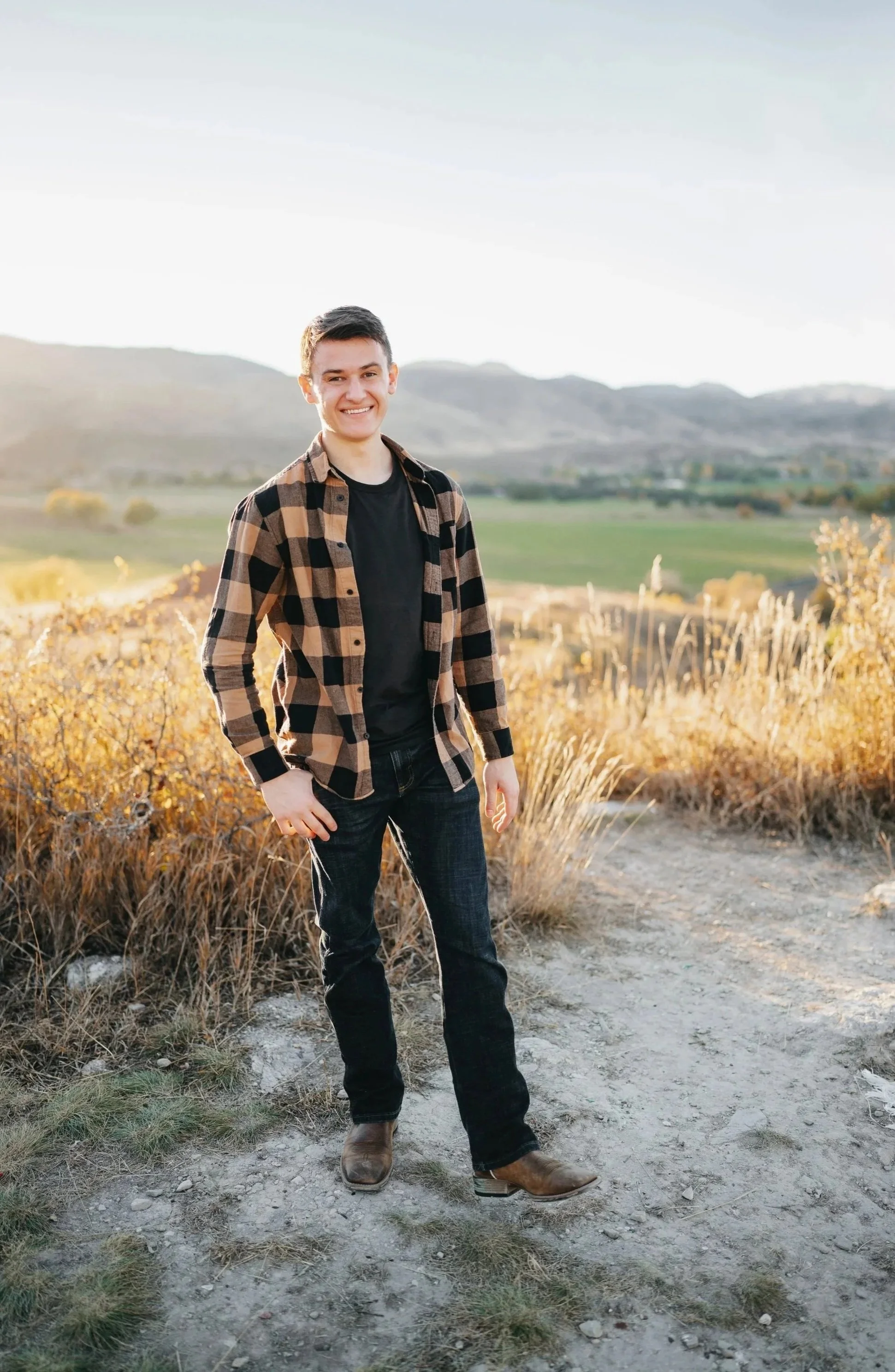 A young man standing outdoors on a dirt path in a rural area with hills and fields in the background. He is smiling, wearing a black T-shirt, black jeans, brown boots, and a brown and black checkered flannel shirt.
