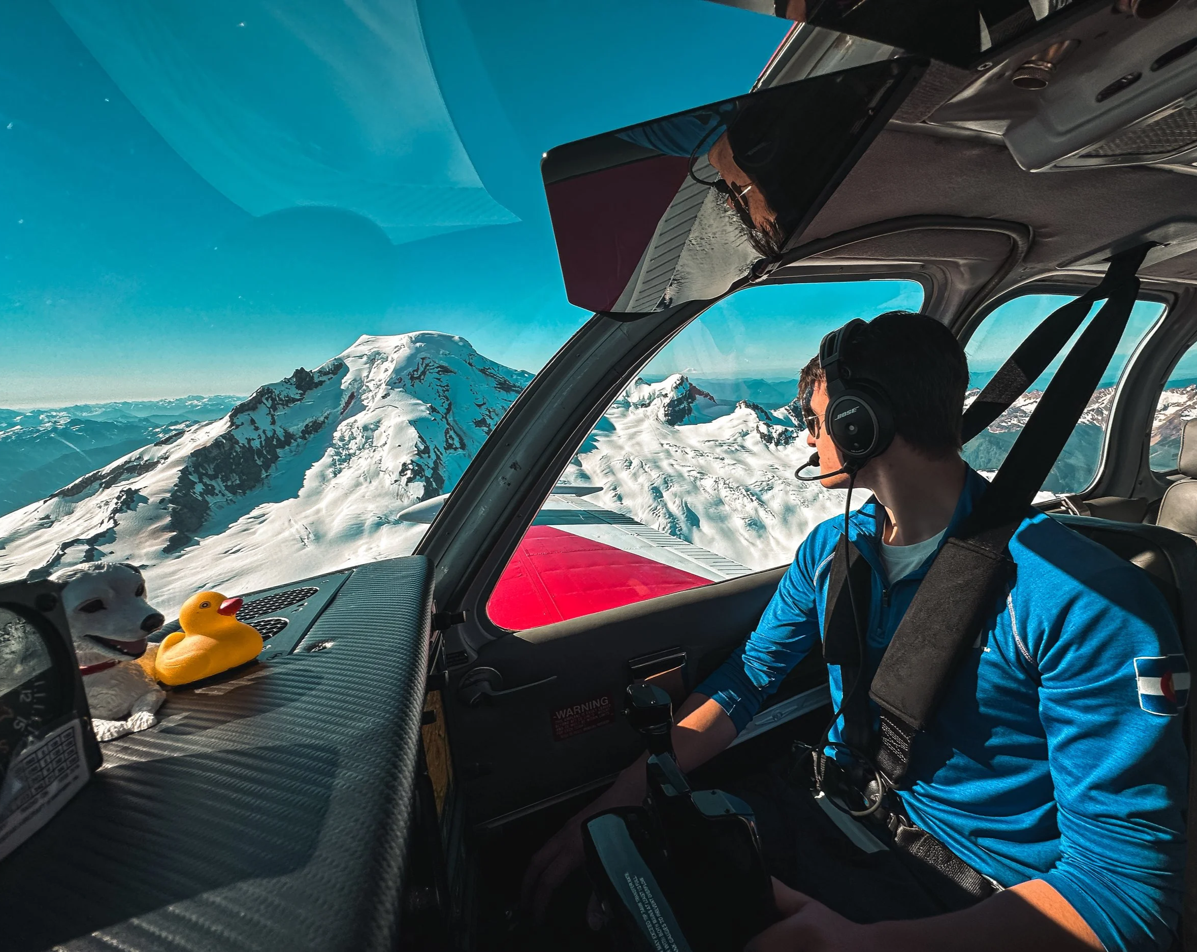 A pilot in a blue jacket flying a small airplane over snow-covered Mount Baker with a clear blue sky in the background.