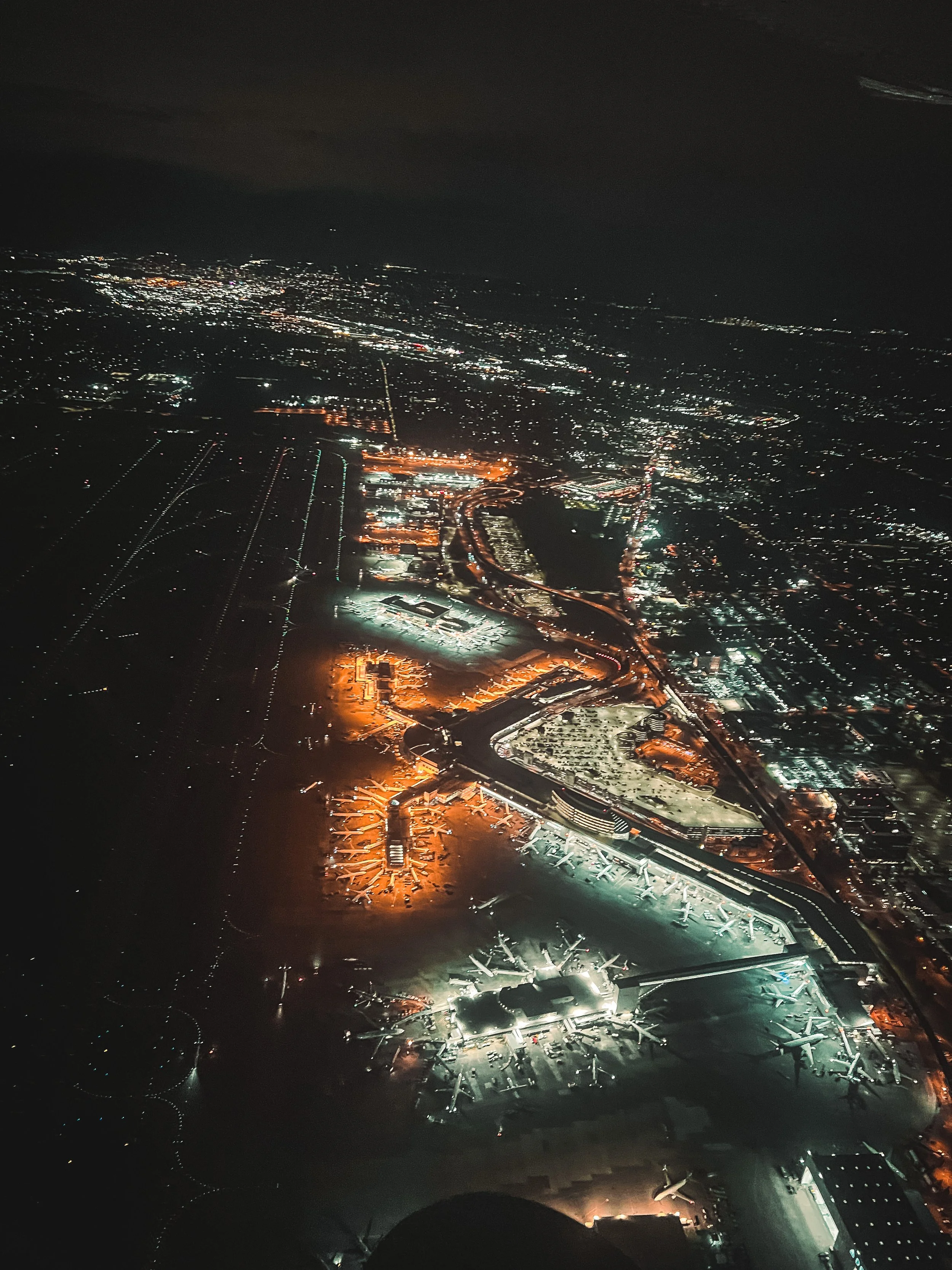 Night aerial view of a large airport (KSEA) with numerous terminals and airplanes, illuminated against the dark sky, with city lights visible in the background.