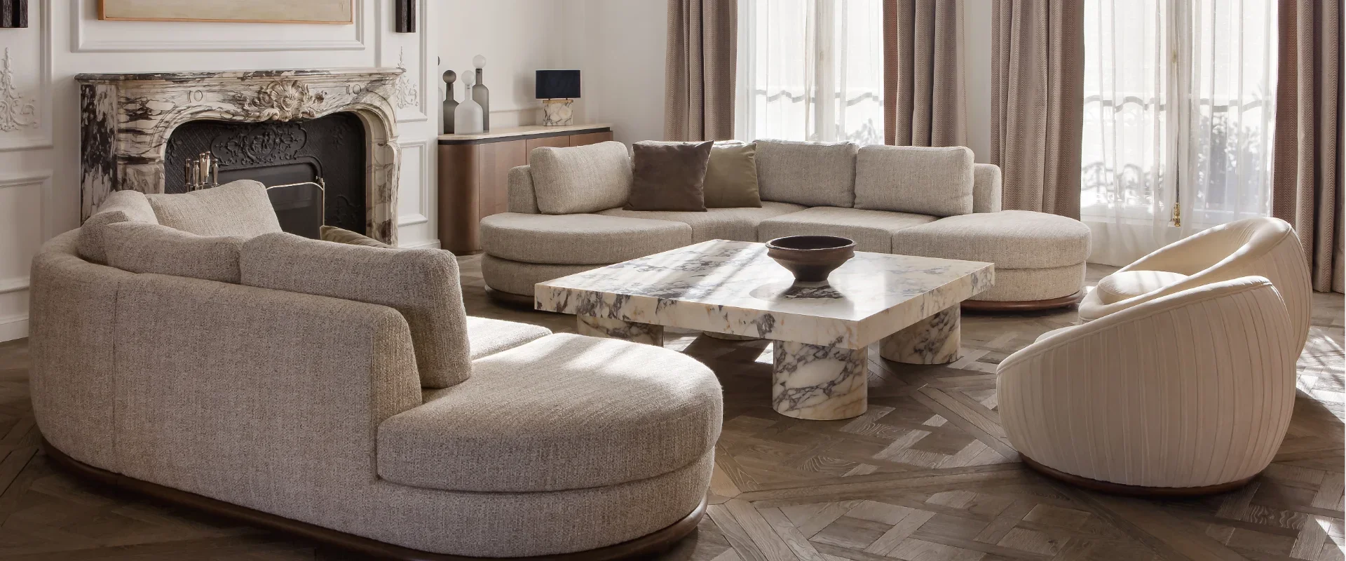 Living room with beige couches, a marble coffee table, and a fireplace with marble surround. Large windows with sheer curtains let in natural light.